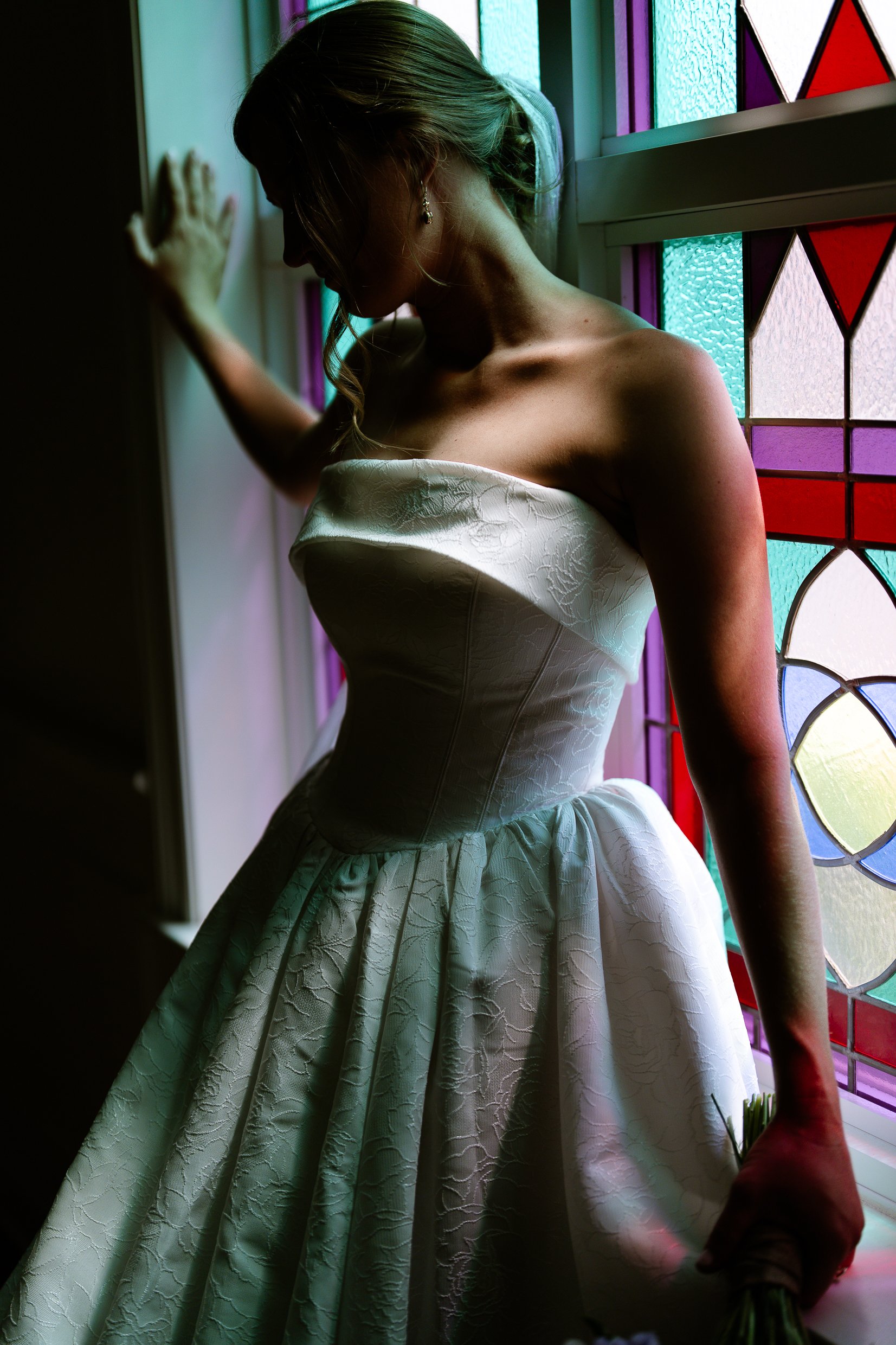 Elizabeth with one hand on a window frame and her face turned away, colored light from the stained glass falling on her shoulder and gown.