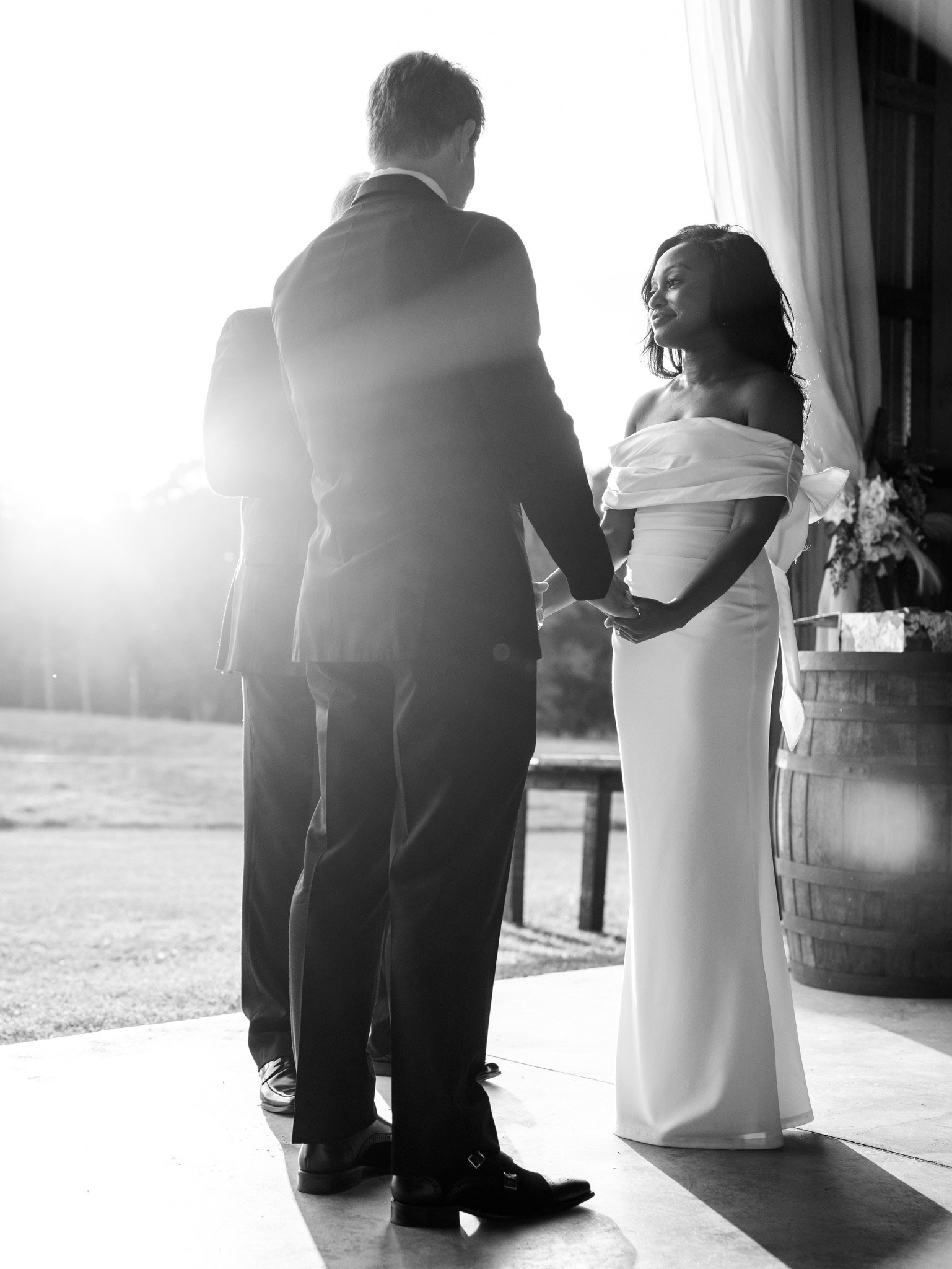 In black and white, Jason and Ashley stand holding hands at the barn opening with the sun behind them.