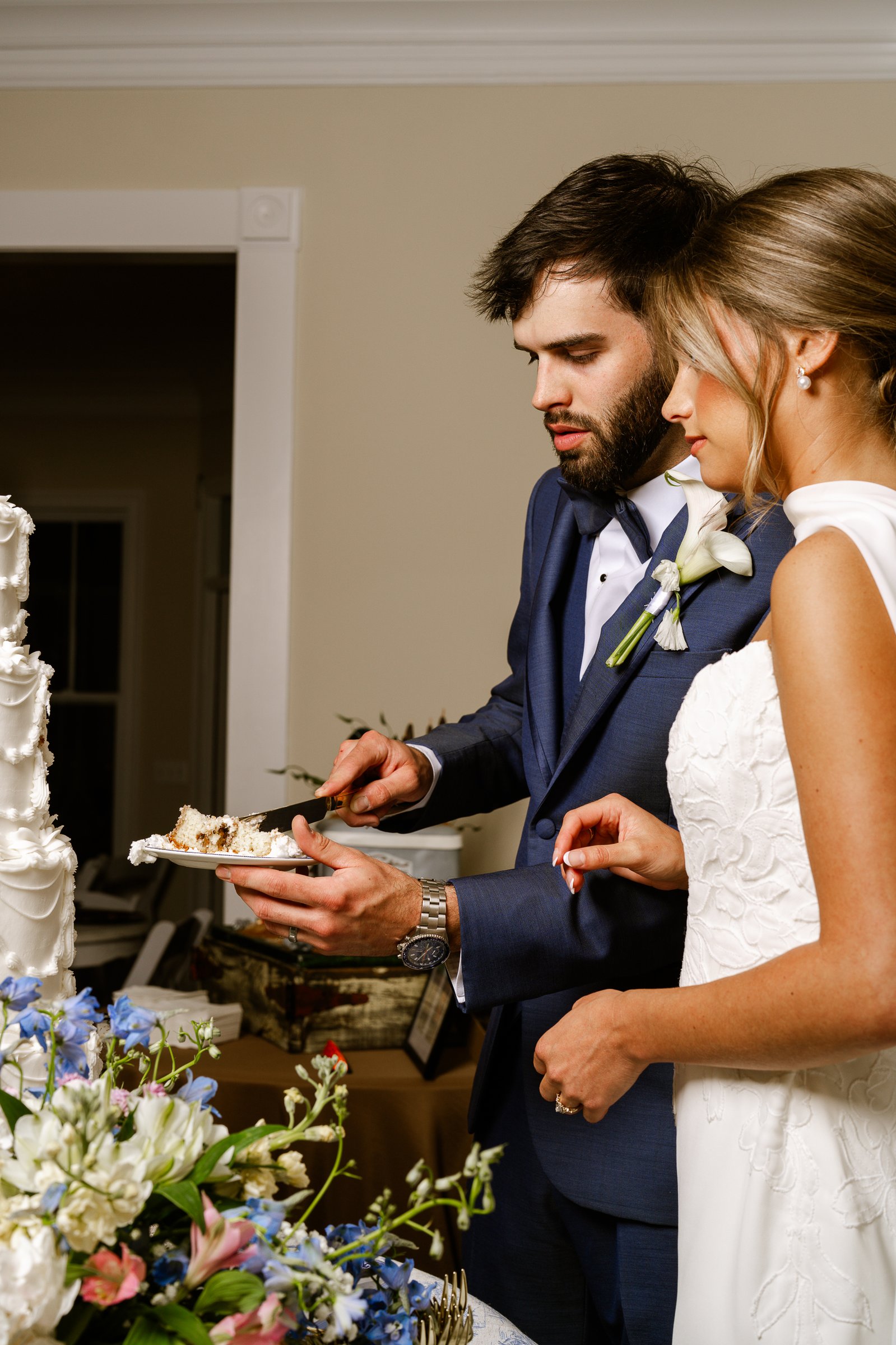 Matthew cuts a slice of cake while Joley stands beside him, with blue and pink flowers in the foreground.
