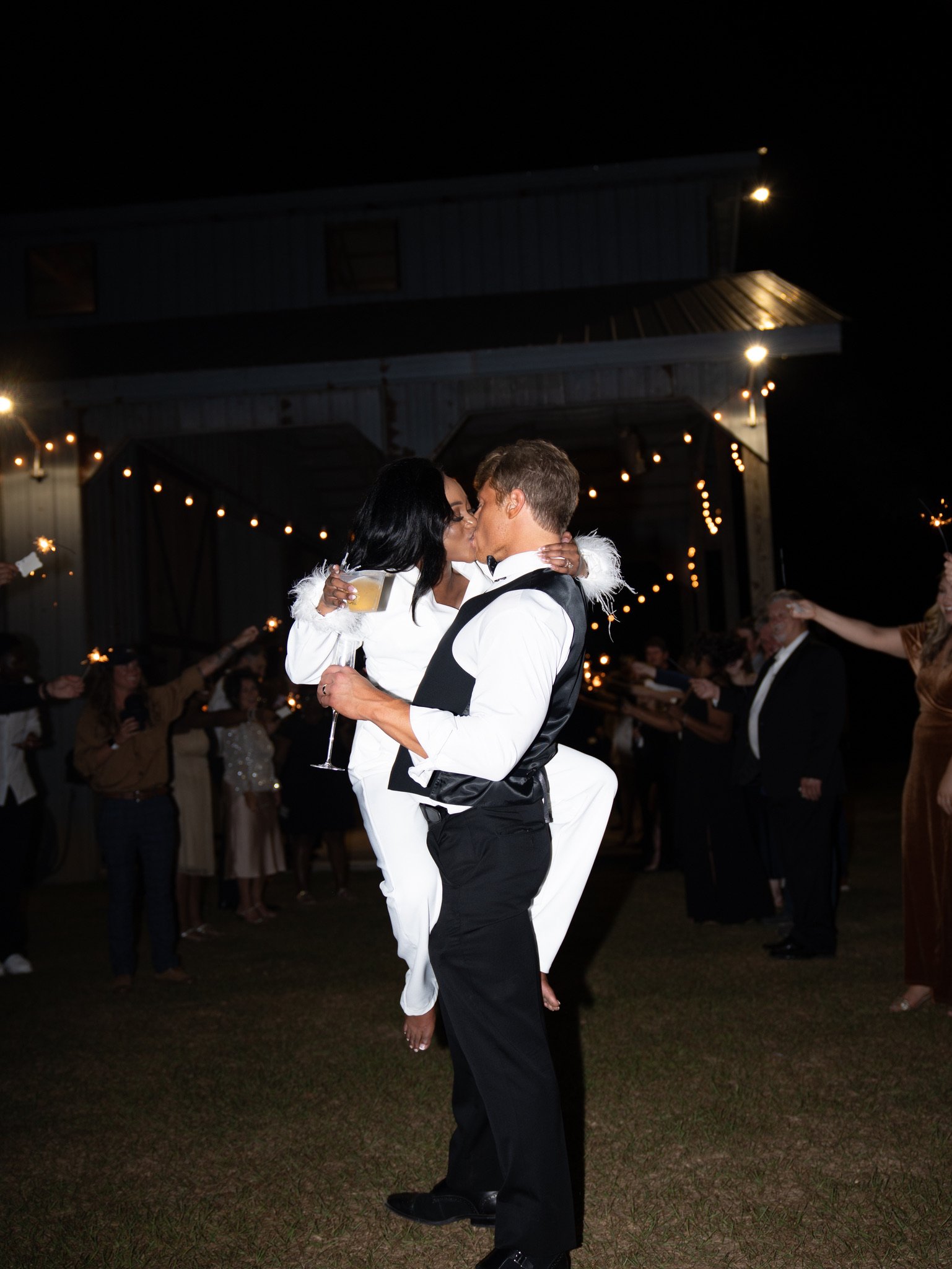 Jason lifts Ashley off the ground as they kiss outside at night, with guests around them.
