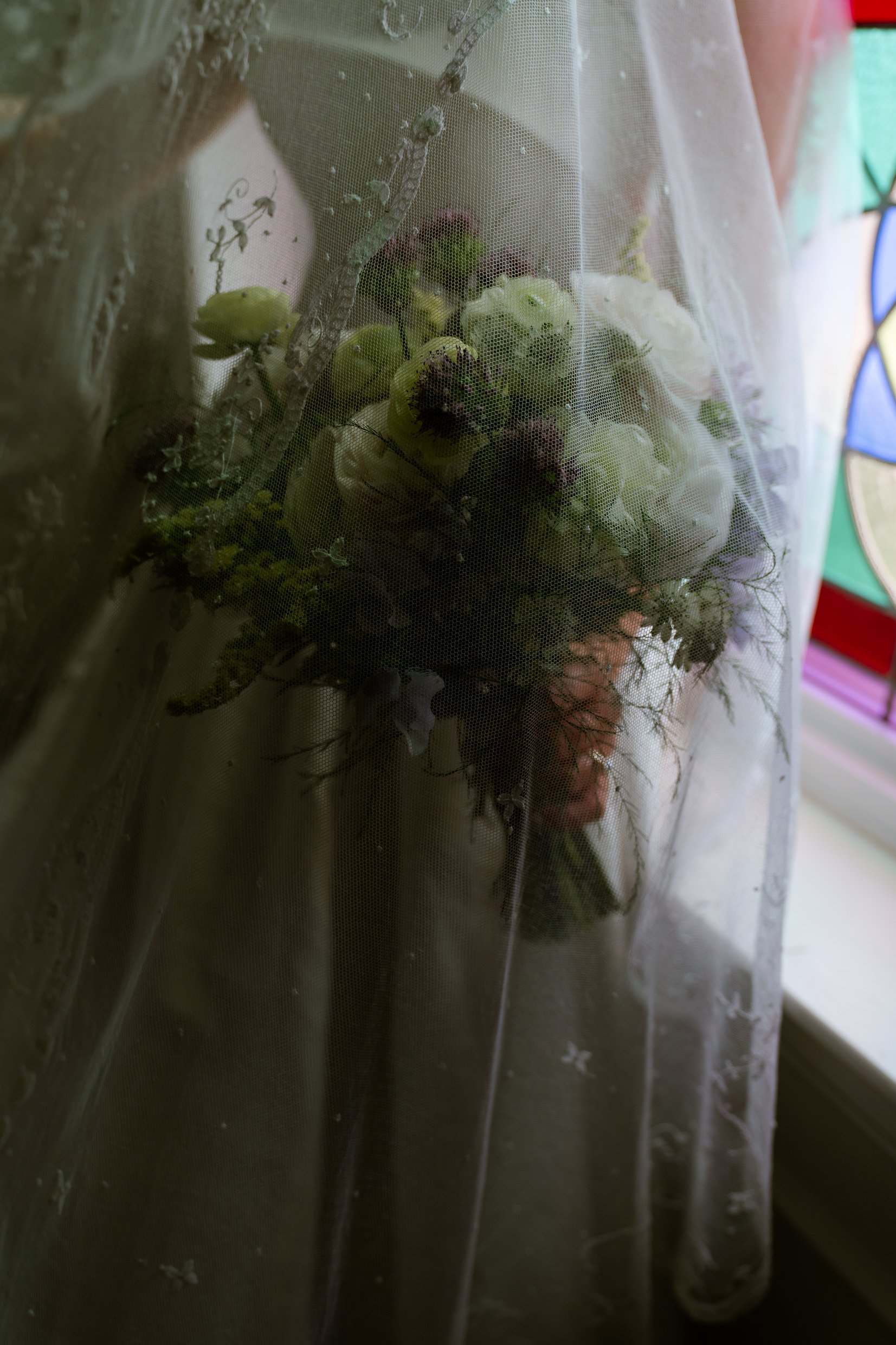 Elizabeth's bouquet of green and white flowers seen through her veil, with a stained glass window beside it.