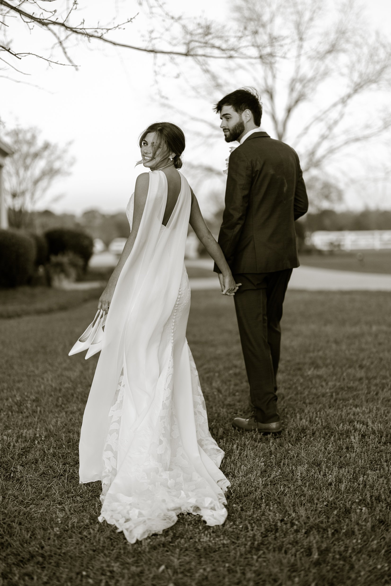 Black and white photo of Joley and Matthew walking away across a lawn holding hands, Joley looking back over her shoulder with her shoes in her hand.