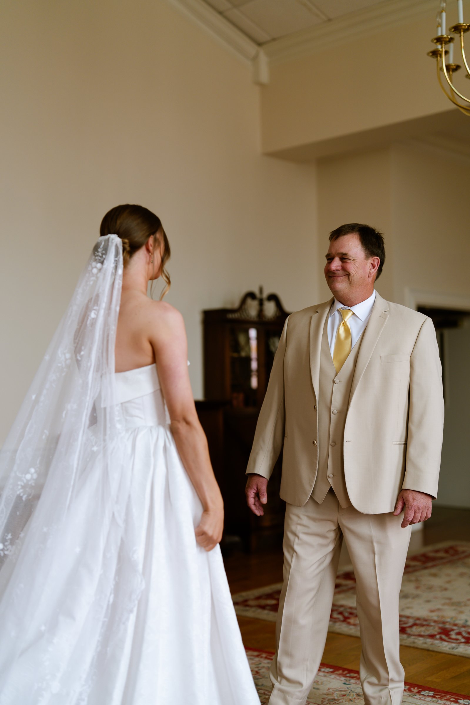 Elizabeth, her veil down her back, stands with her back to the camera as her father in a tan suit smiles at her across a bright room.