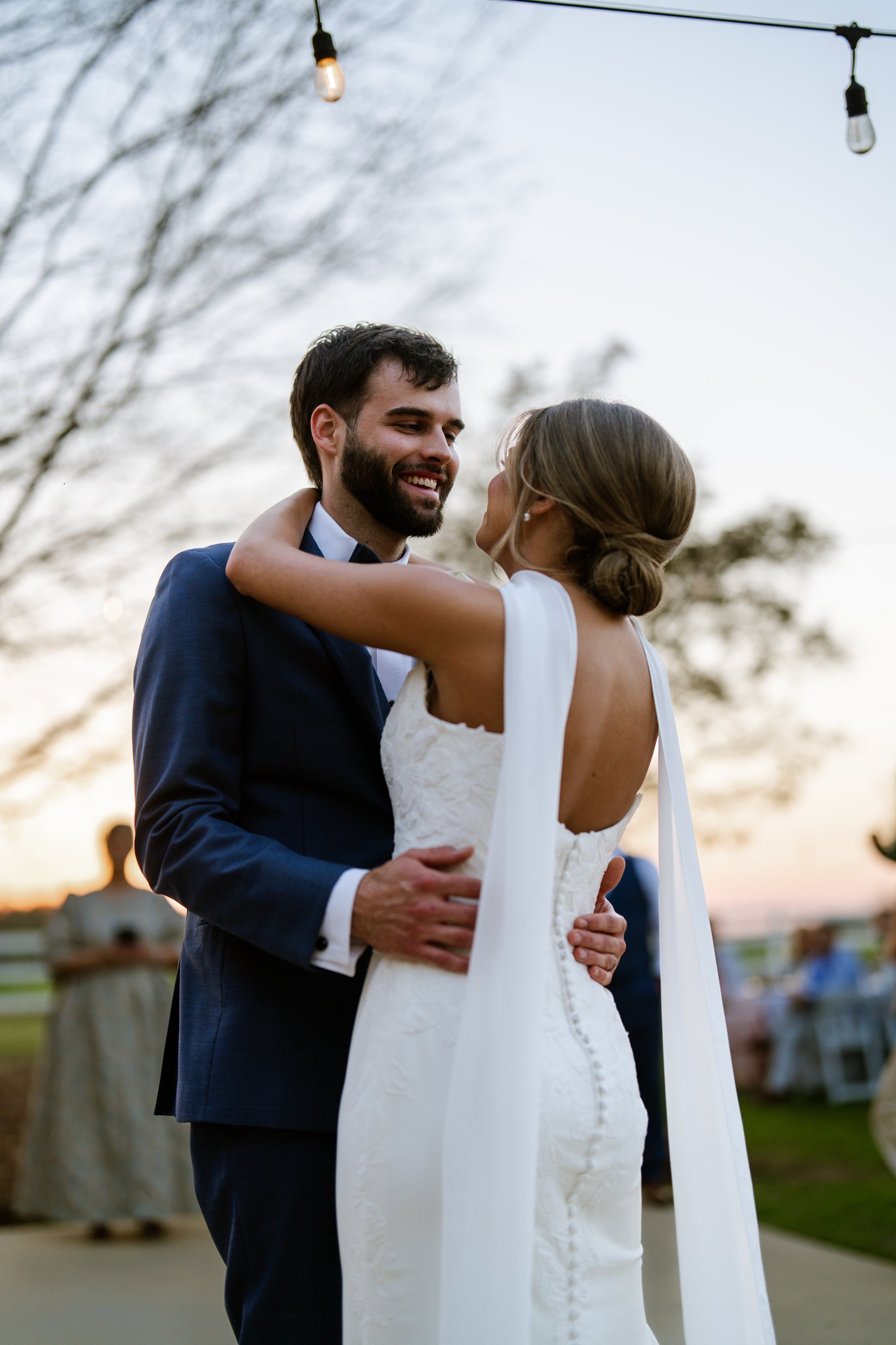 Joley and Matthew smile at each other in an embrace at dusk, with string lights and bare branches above them.