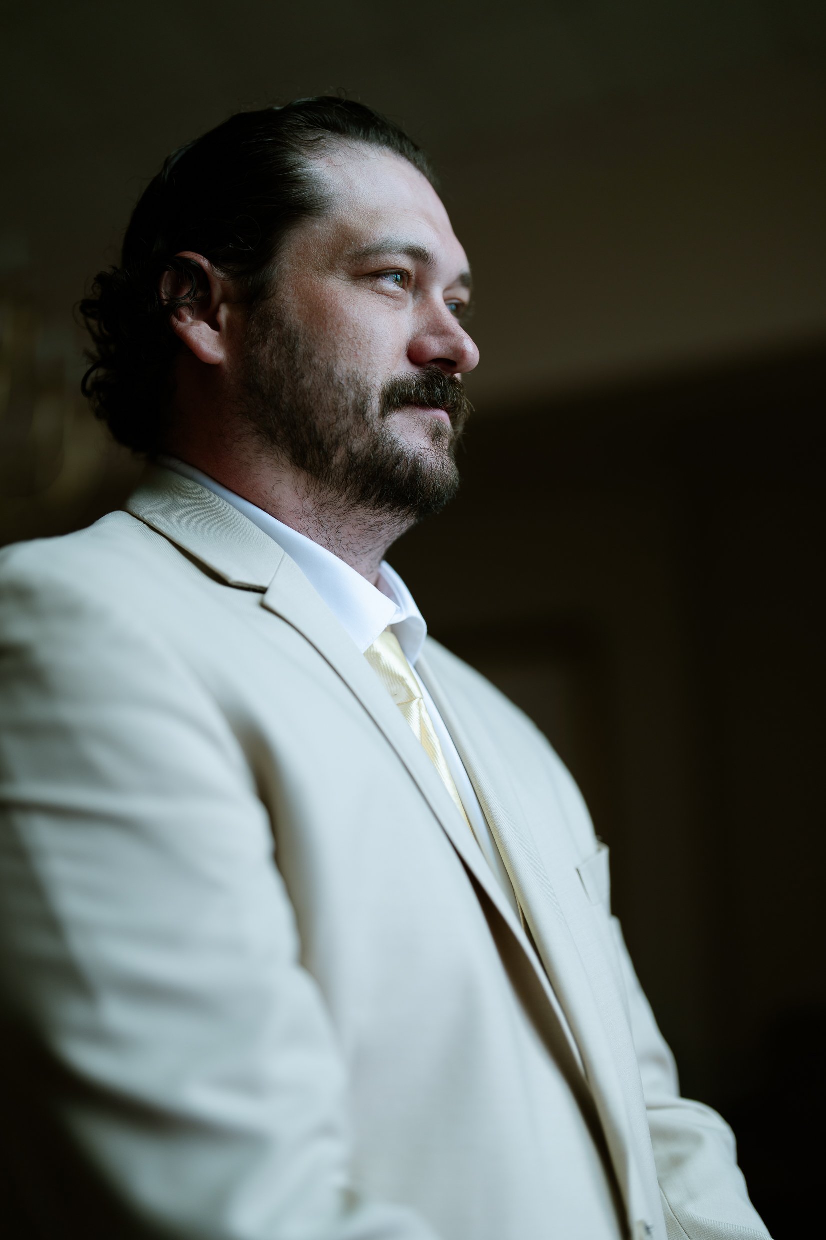 Dustin in a tan suit and yellow tie looks off to the side in the dim light of the church.