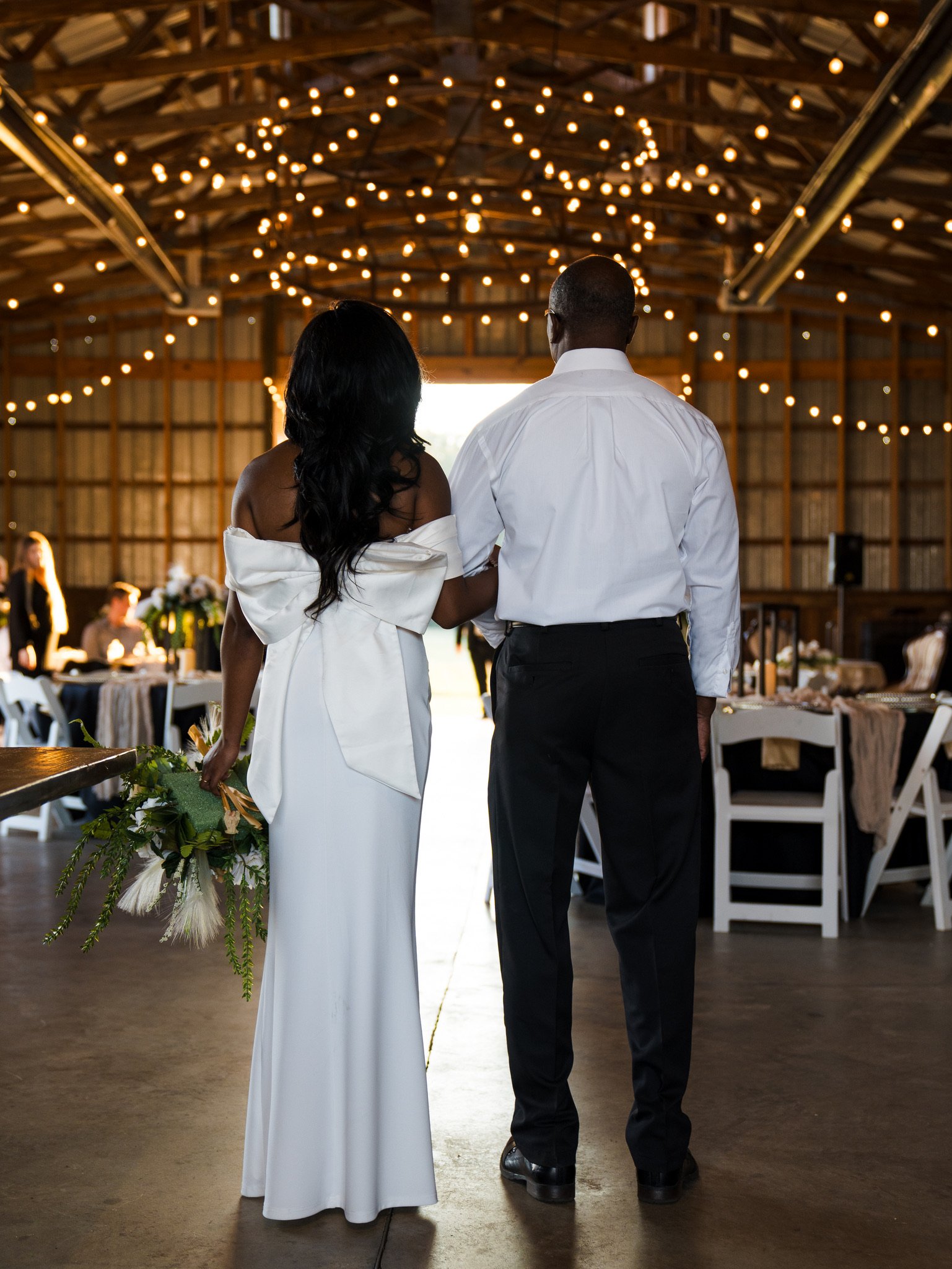 Ashley in a white dress with a large bow stands arm in arm with a man in a white shirt, facing the barn doorway under string lights.
