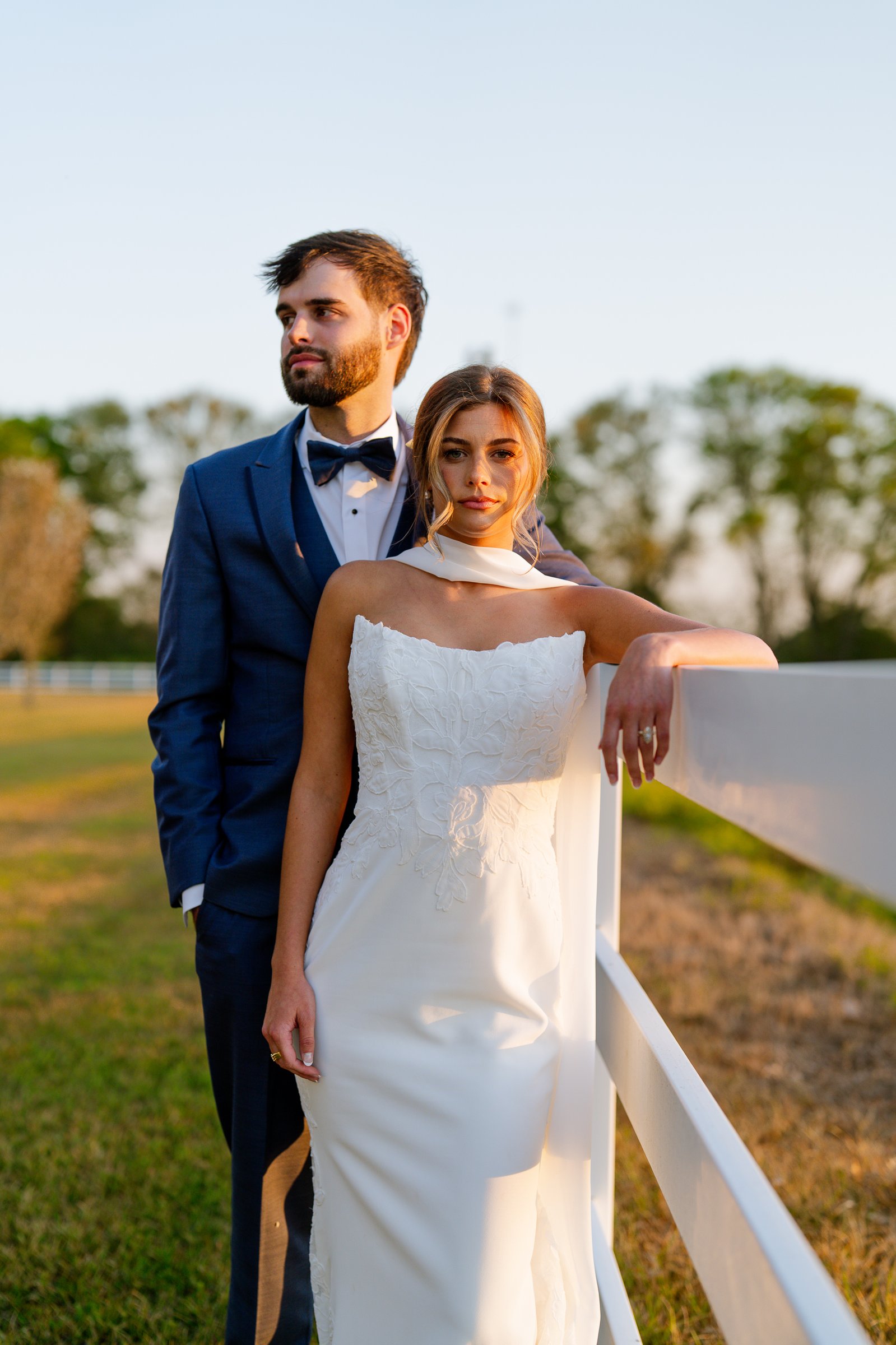 Joley and Matthew stand together at a white fence in warm evening light, Matthew in a blue suit and bow tie and Joley in a white strapless dress looking at the camera.