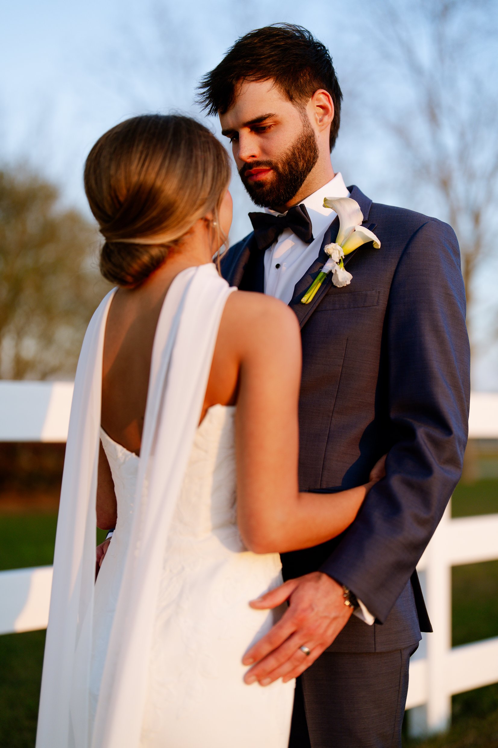 Joley, with her back to the camera and a sheer wrap down her back, embraces Matthew in front of a white fence.