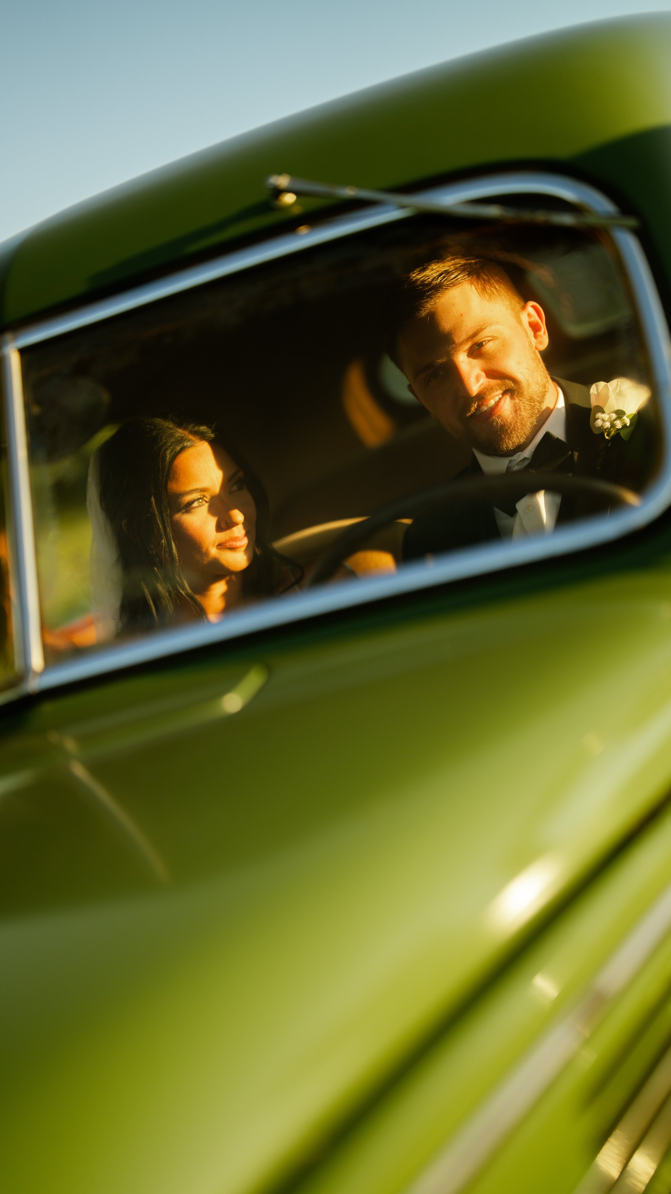 Kenadee and Corey seen through the window of a green vintage car, looking at each other.