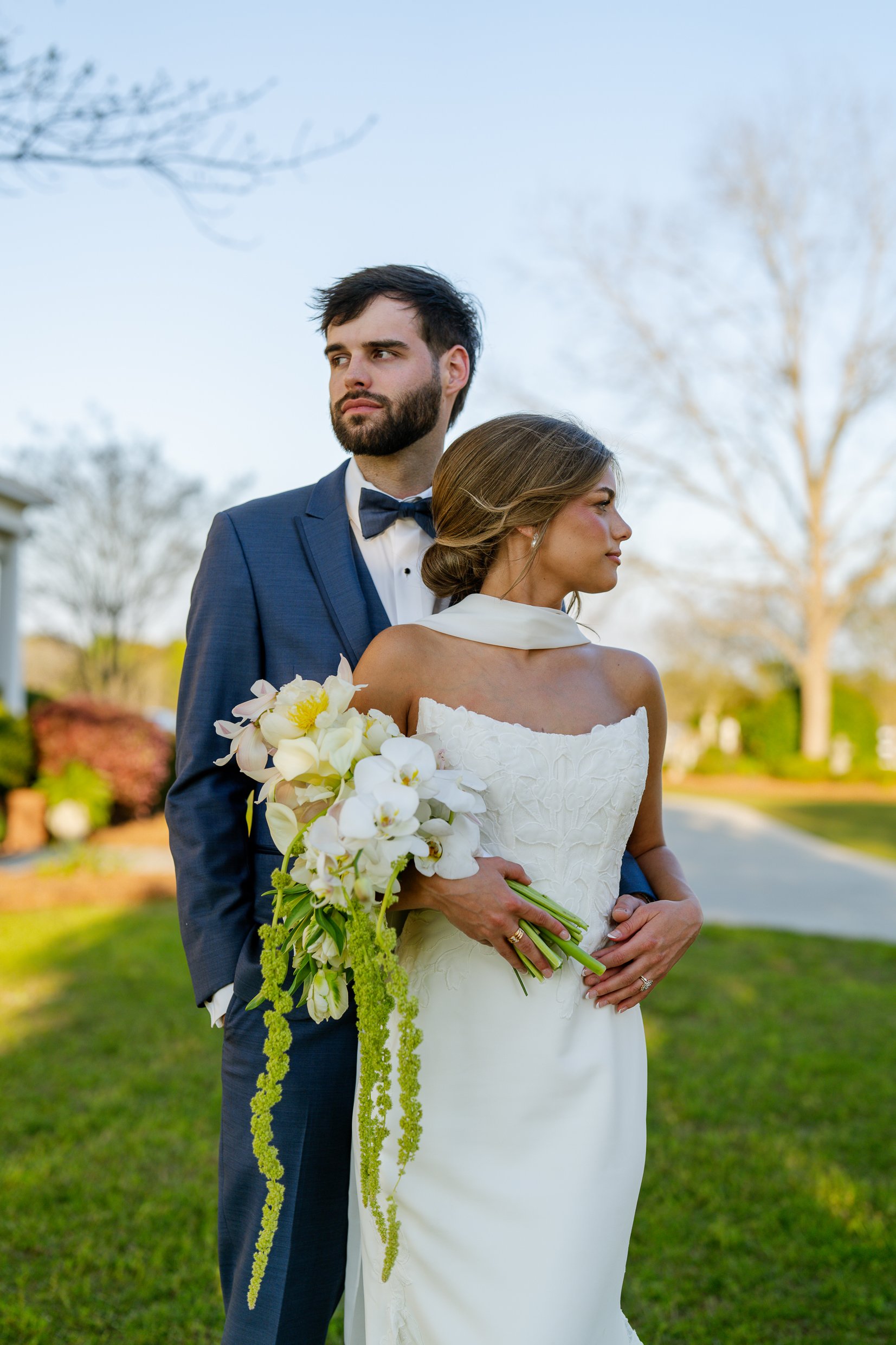 Matthew stands behind Joley, who holds a white and yellow bouquet with trailing greenery, both looking off to the side across a lawn.