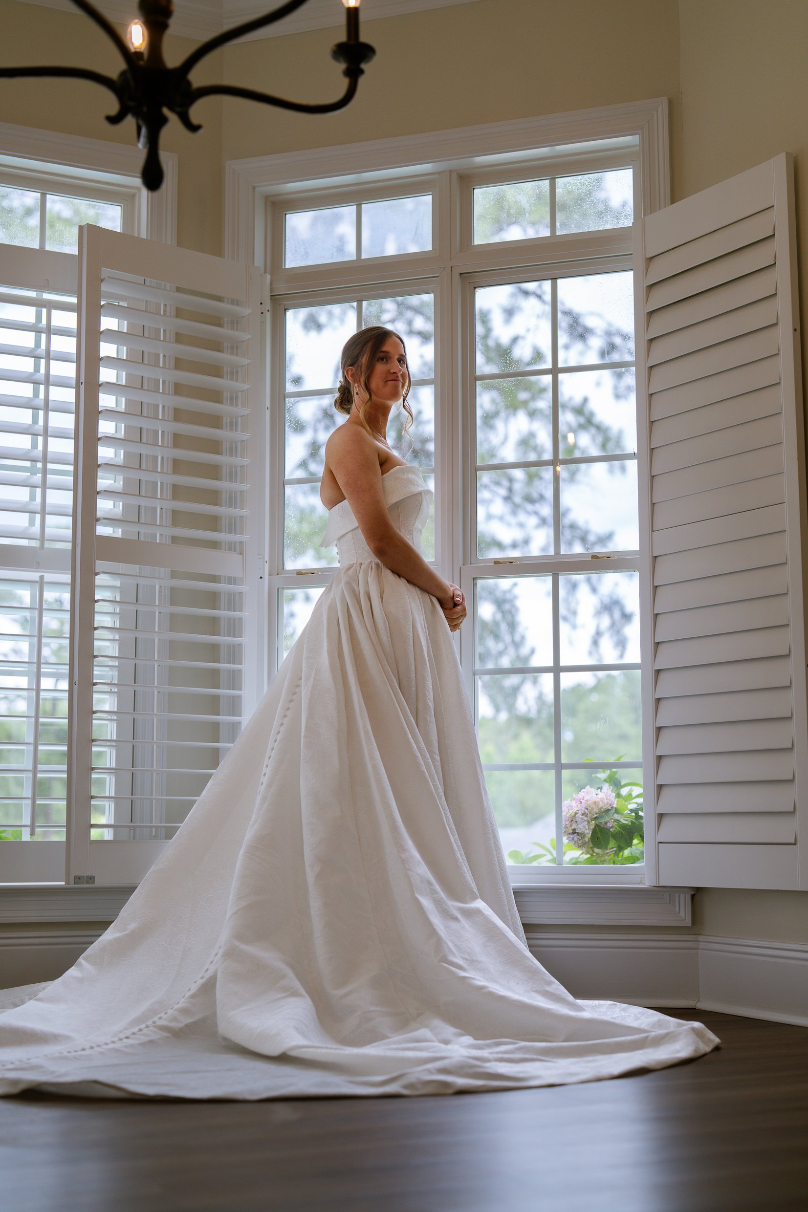 Elizabeth stands sideways in a bright room with tall shuttered windows, her long white train spread across the wood floor.