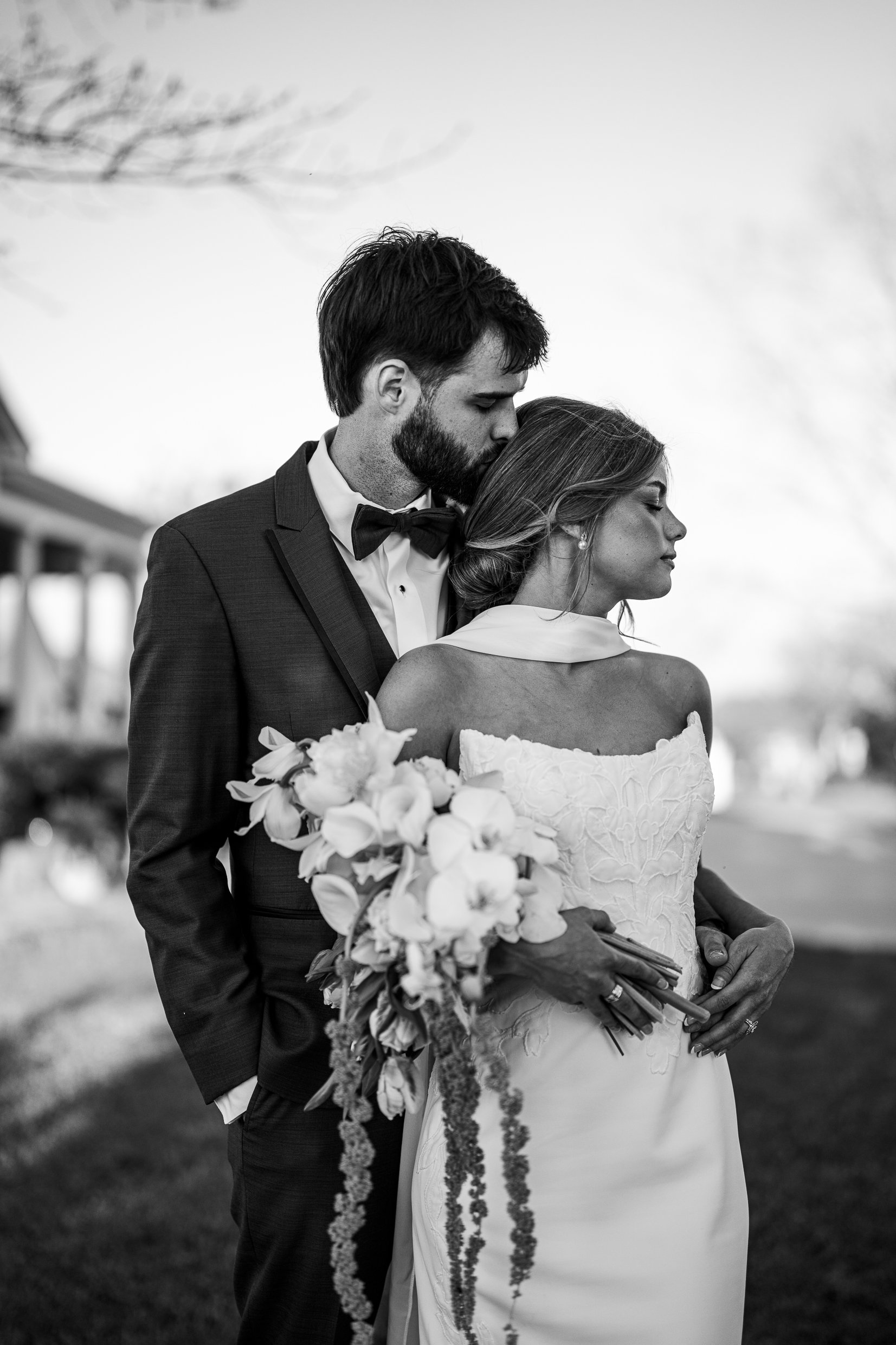 Black and white photo of Matthew kissing the top of Joley's head while she holds a large bouquet.