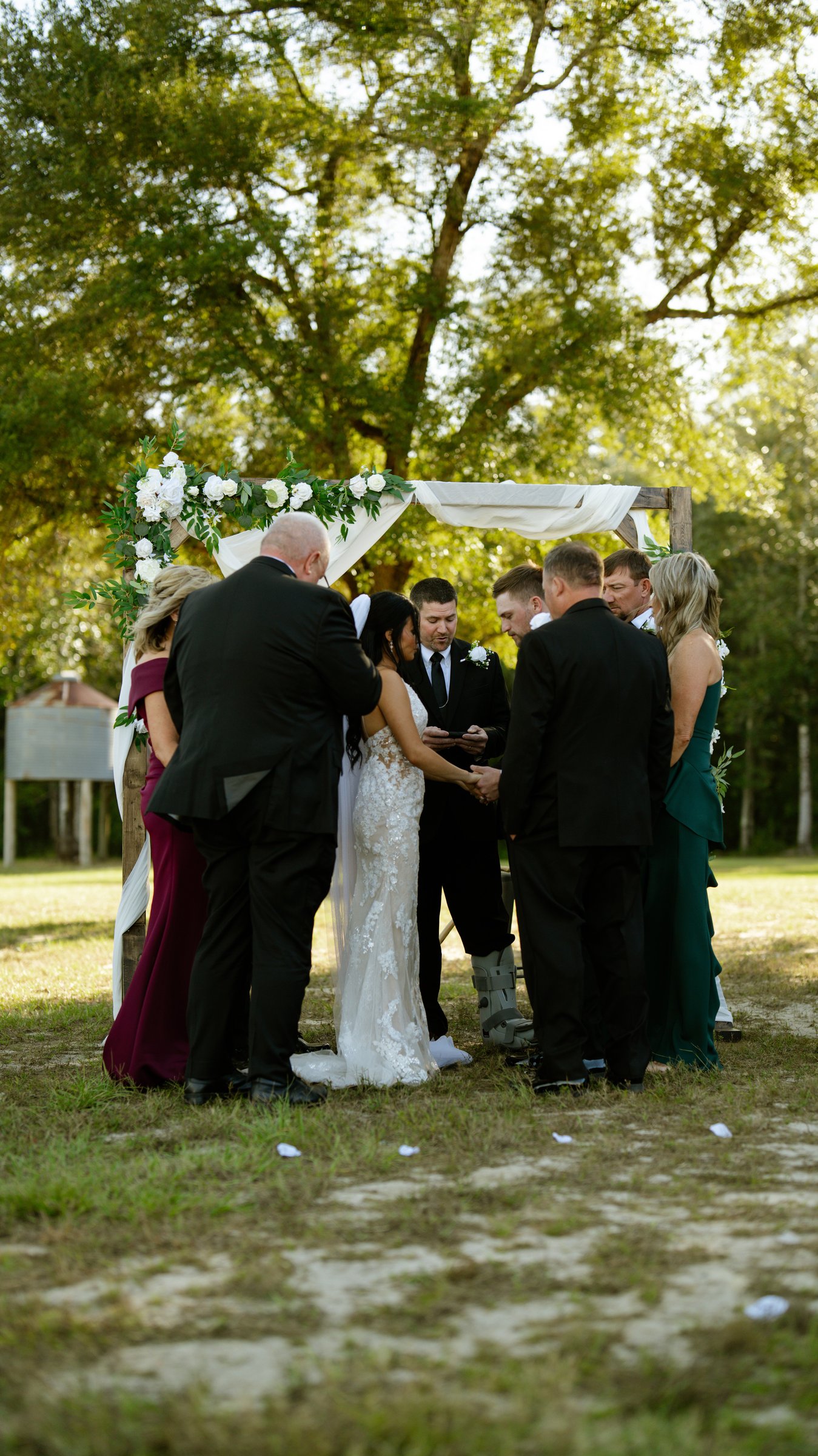 Kenadee and Corey standing under a flower arch at the ceremony, with guests around them under big trees.