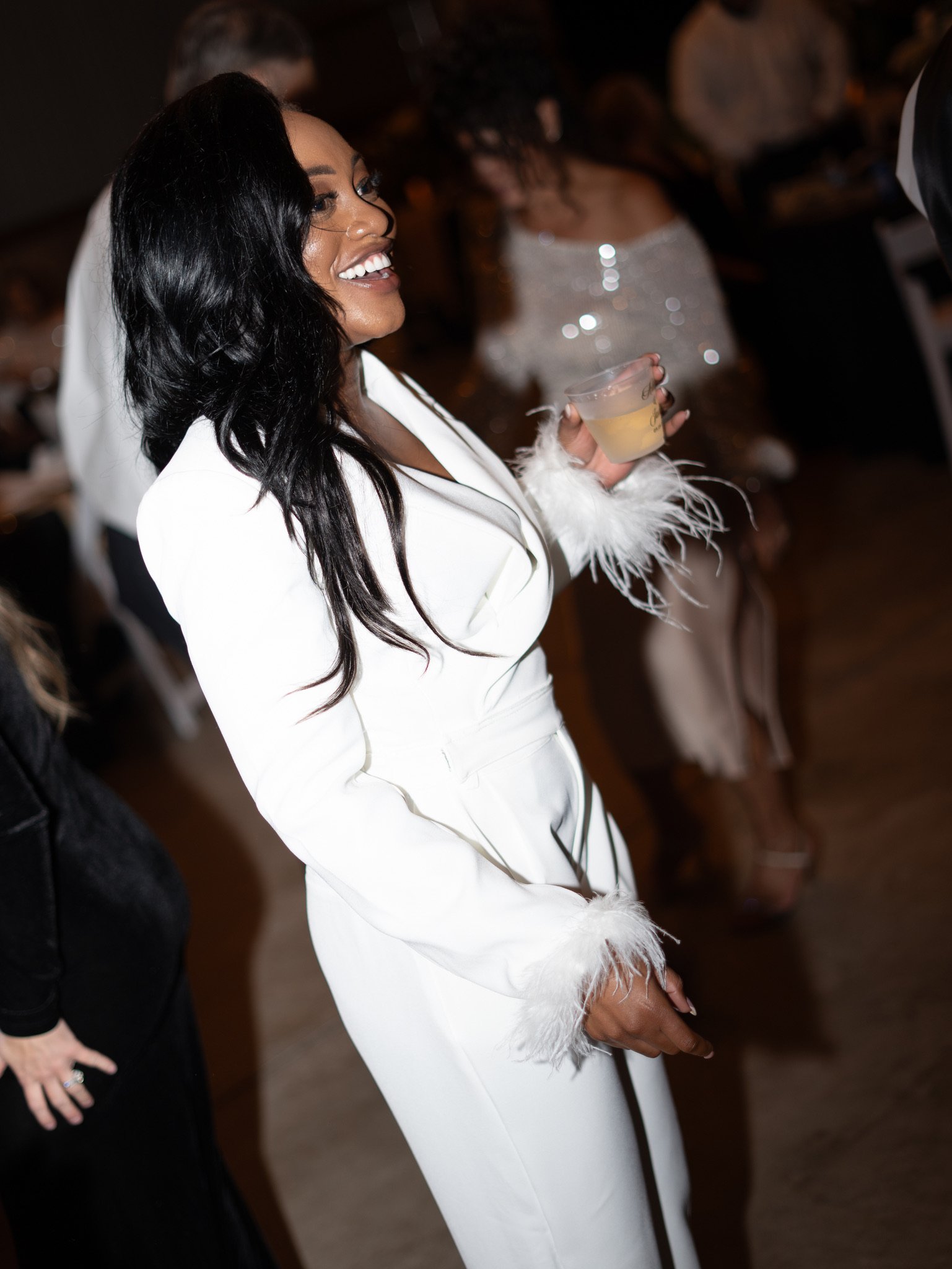 A woman in a white suit with feathered cuffs laughs and holds a drink on a crowded dance floor.