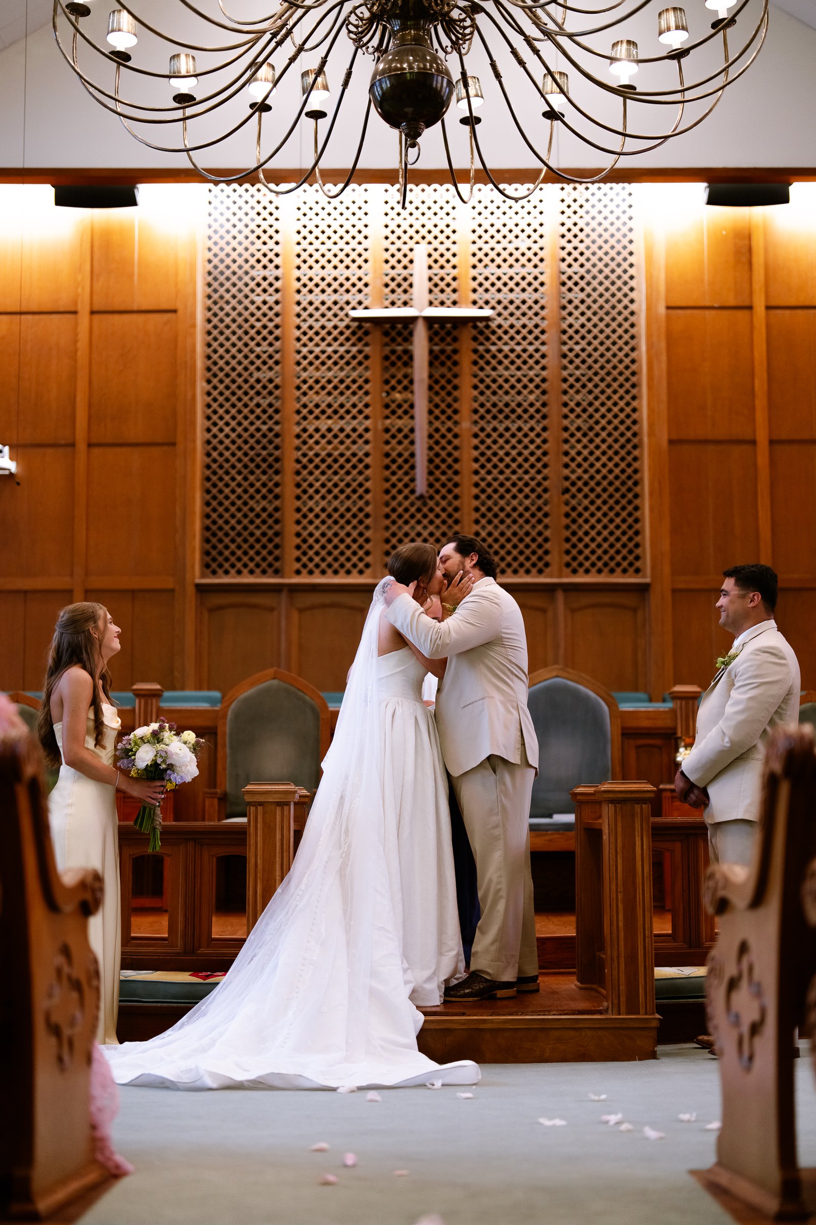 Elizabeth and Dustin kiss at the front of the church under a wooden cross, with a chandelier above.