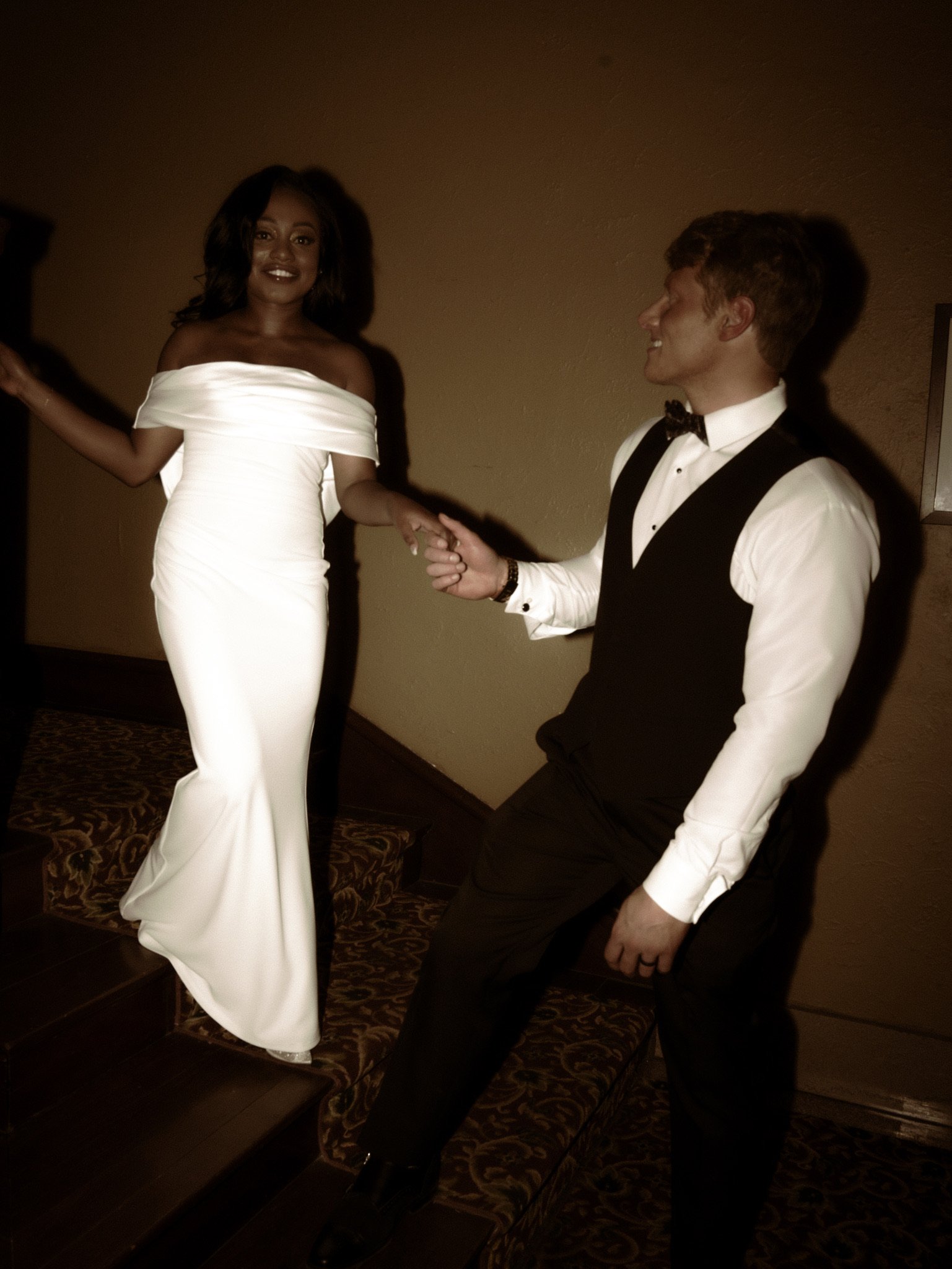 Ashley in a white dress and Jason in a vest walk down a carpeted staircase holding hands, in a warm brown tone.