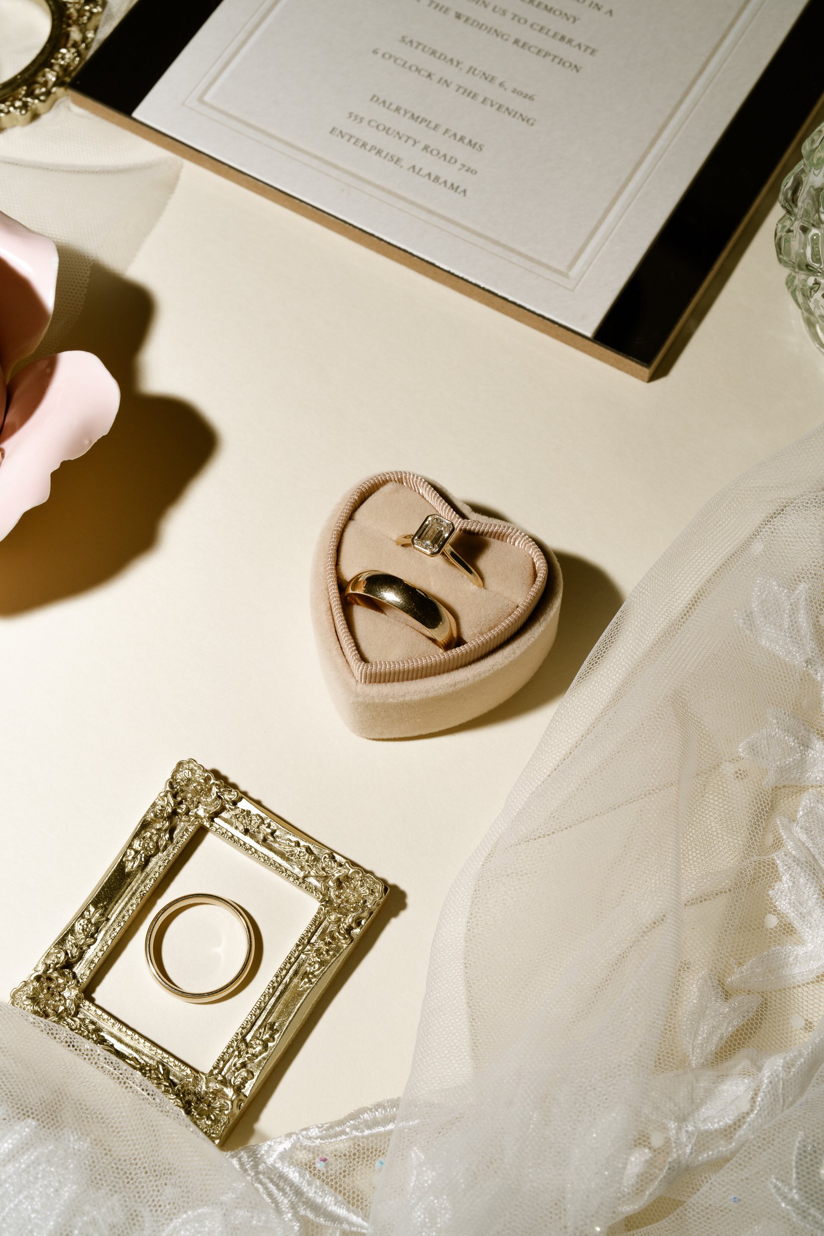 Two gold rings in a blush heart-shaped box beside a small gold frame holding another ring, with the invitation above.