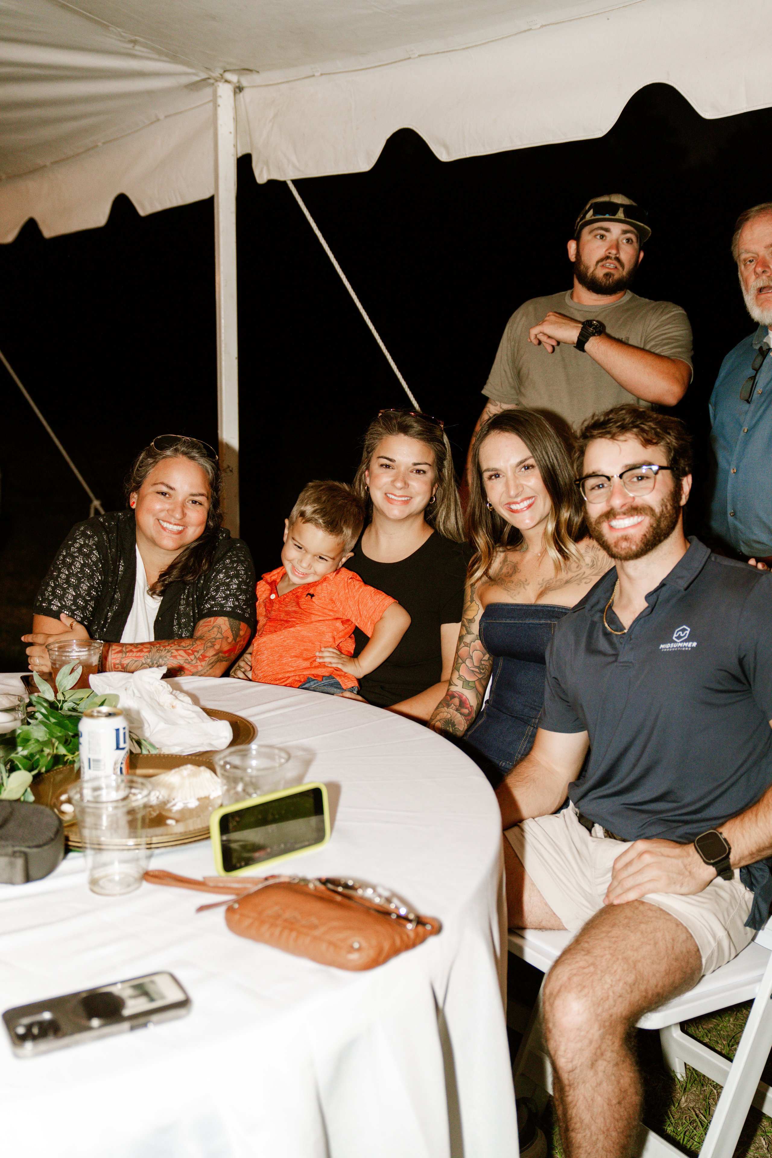 Guests sitting around a round table under a tent at night, smiling at the camera.