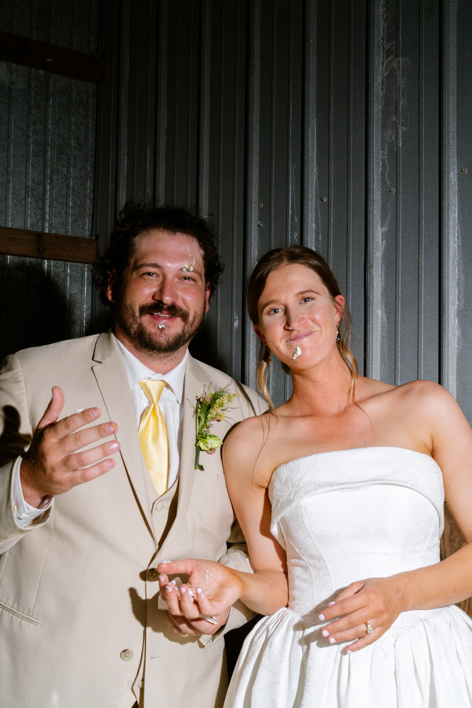 Dustin and Elizabeth look at the camera and laugh with cake on their faces, in front of the barn wall.