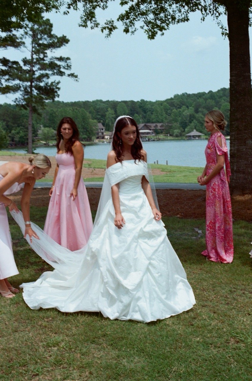 Brooklyn and her bridesmaids gather beside the lake at New Water Farms, with her dress train spread out, photographed on film.