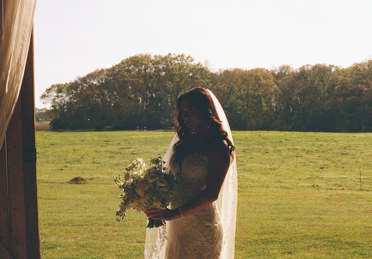 Raley holding her bouquet in a sunlit doorway with the fields of Dalrymple Farms behind her, photographed on film.