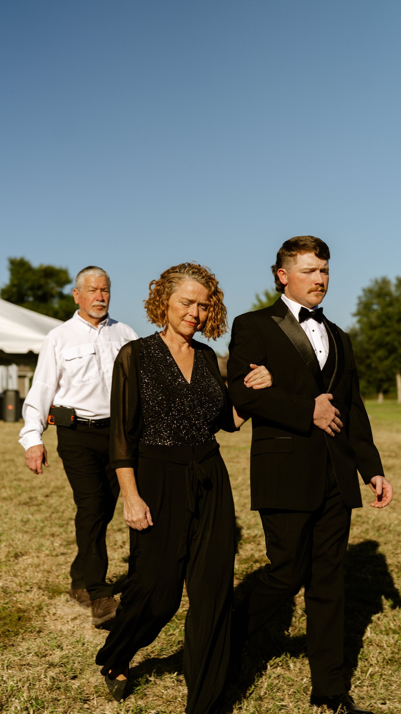 A man in a black tuxedo walks across a dry field arm in arm with a woman in black, with another man behind them.