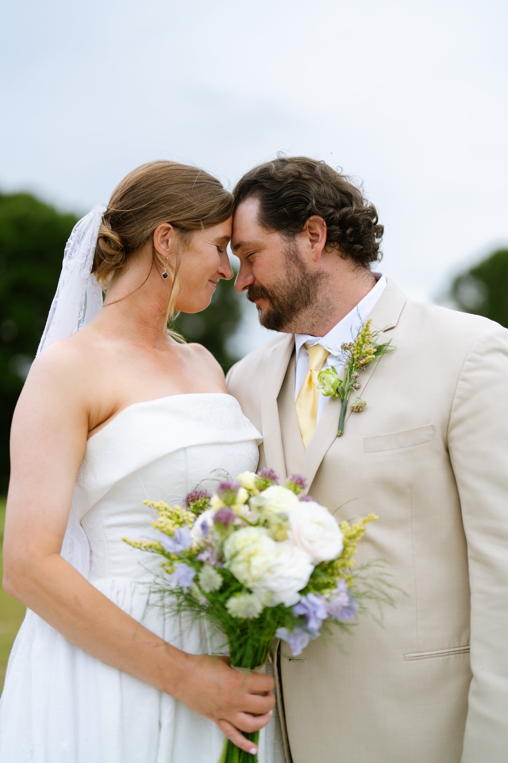 Elizabeth and Dustin touch foreheads outdoors under a cloudy sky, her bouquet of white and lavender flowers in her hand.