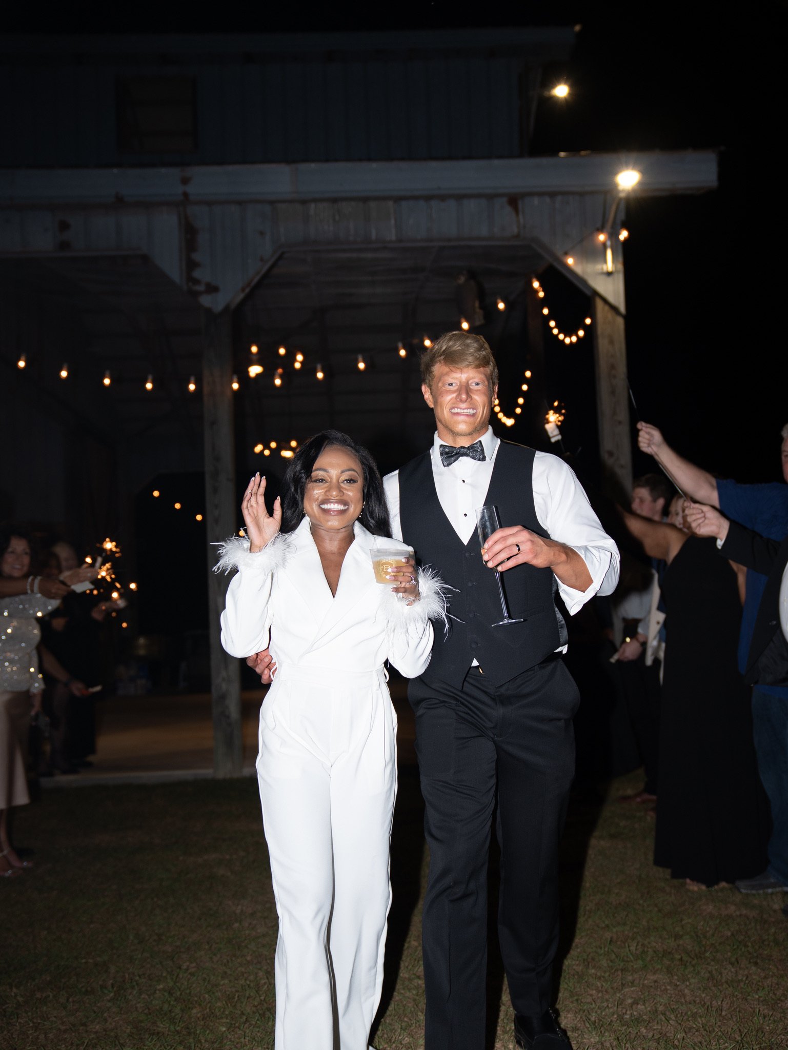 Ashley waves and Jason holds a glass as they walk through a line of guests at night under string lights.