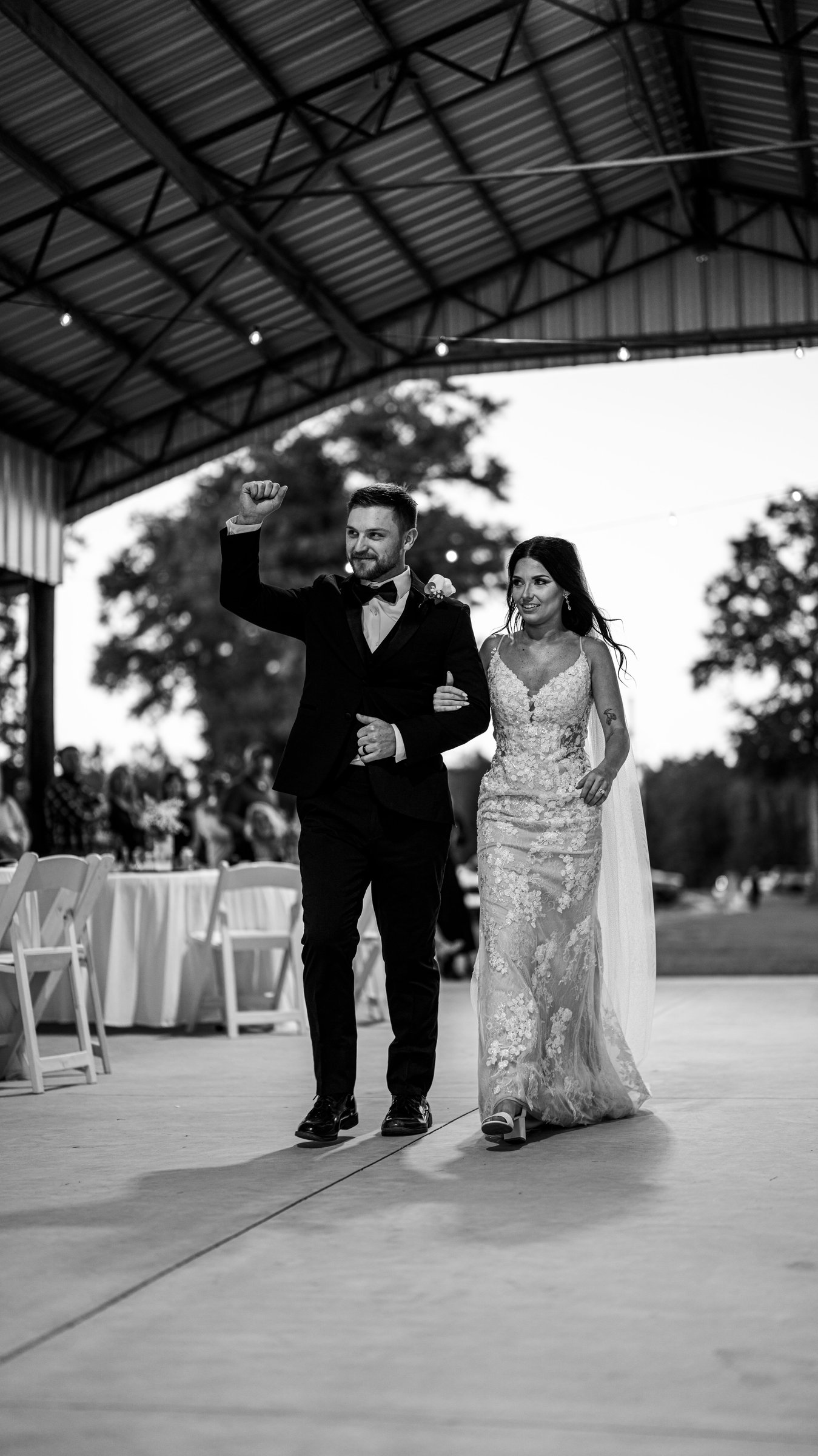 Corey with a fist raised and Kenadee in her dress walking onto the dance floor under a metal-roofed pavilion.