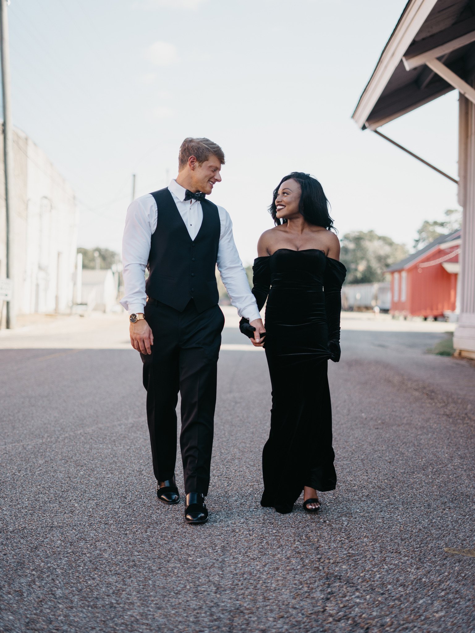 Jason and Ashley walk down the street side by side holding hands and looking at each other, with a red building behind them.