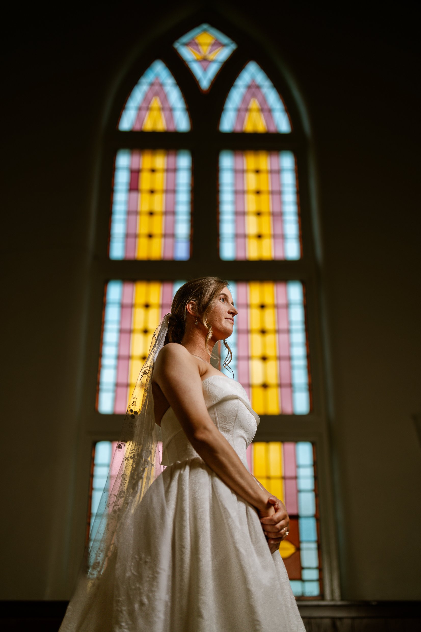 Elizabeth in profile in front of a tall stained glass window, her veil trailing over her shoulder as she looks into the light.