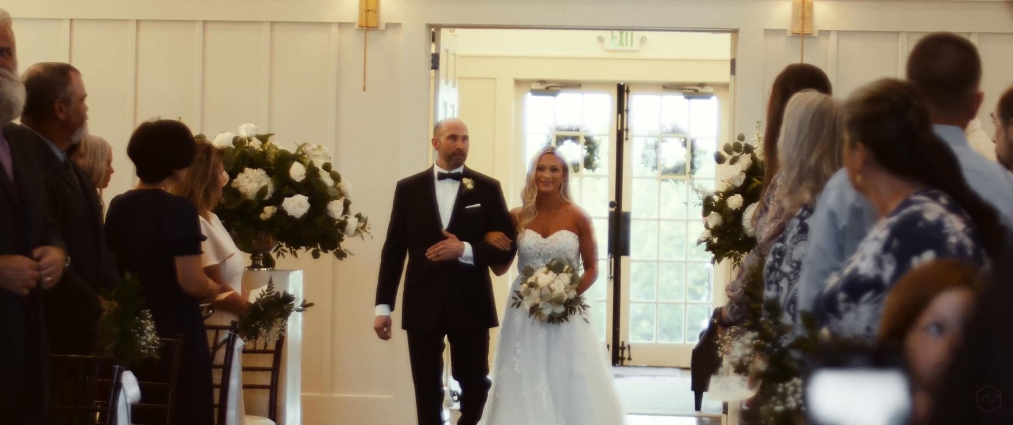 Madalyn walks down the aisle with her dad as their wedding guests watch. Still from their Midsummer Productions wedding film.