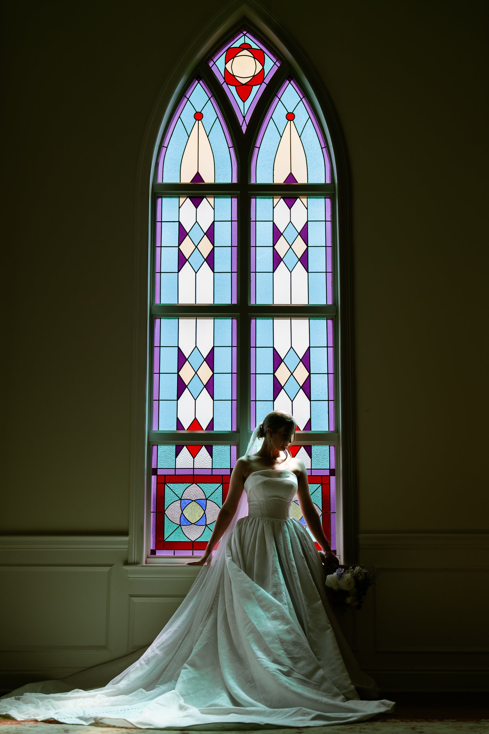 Elizabeth at the base of a tall arched stained glass window, her gown and long train spread across the floor, a bouquet in her hand.