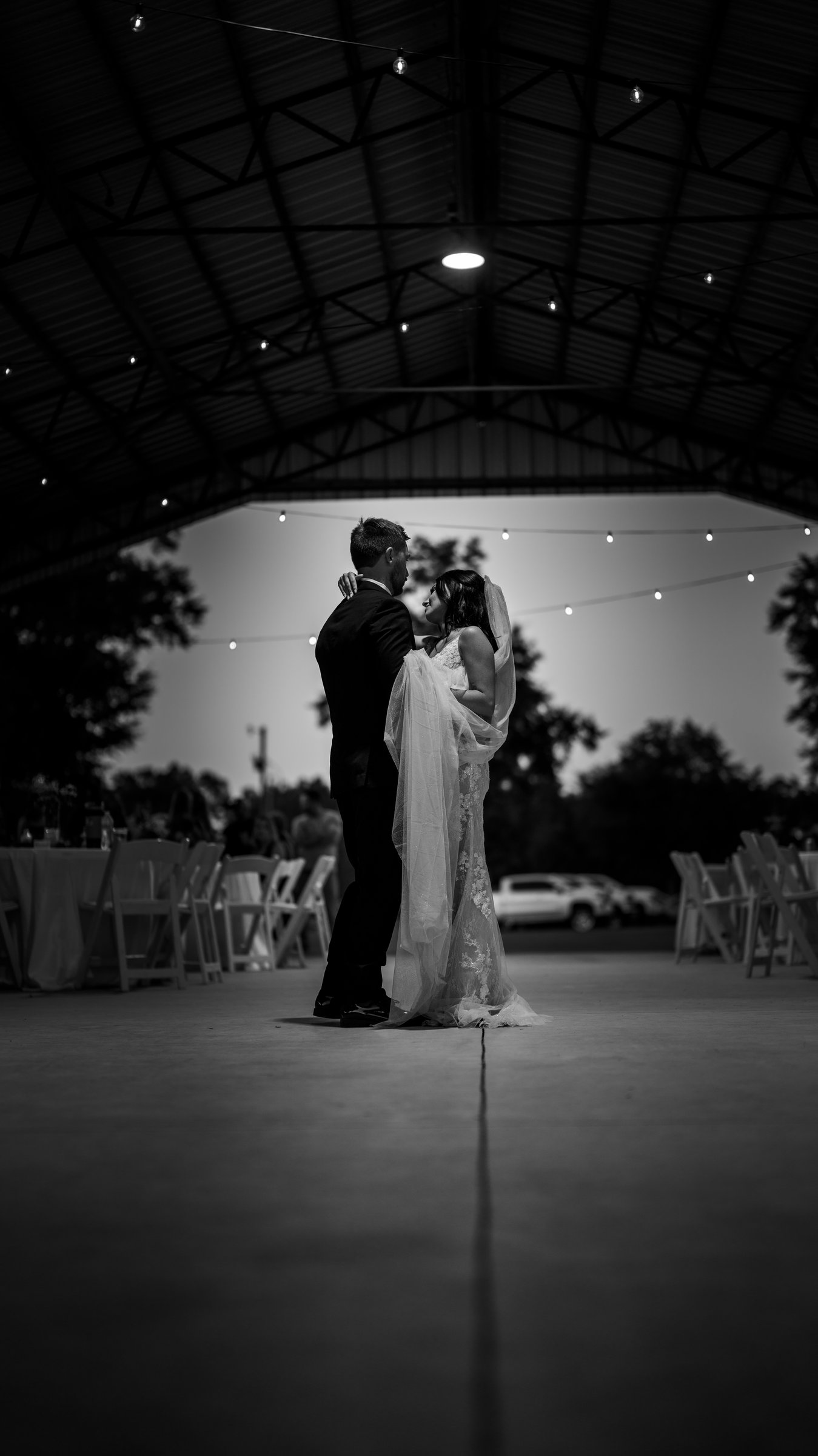 In black and white, Kenadee and Corey dancing in the distance on a wide floor under a dark pavilion roof.