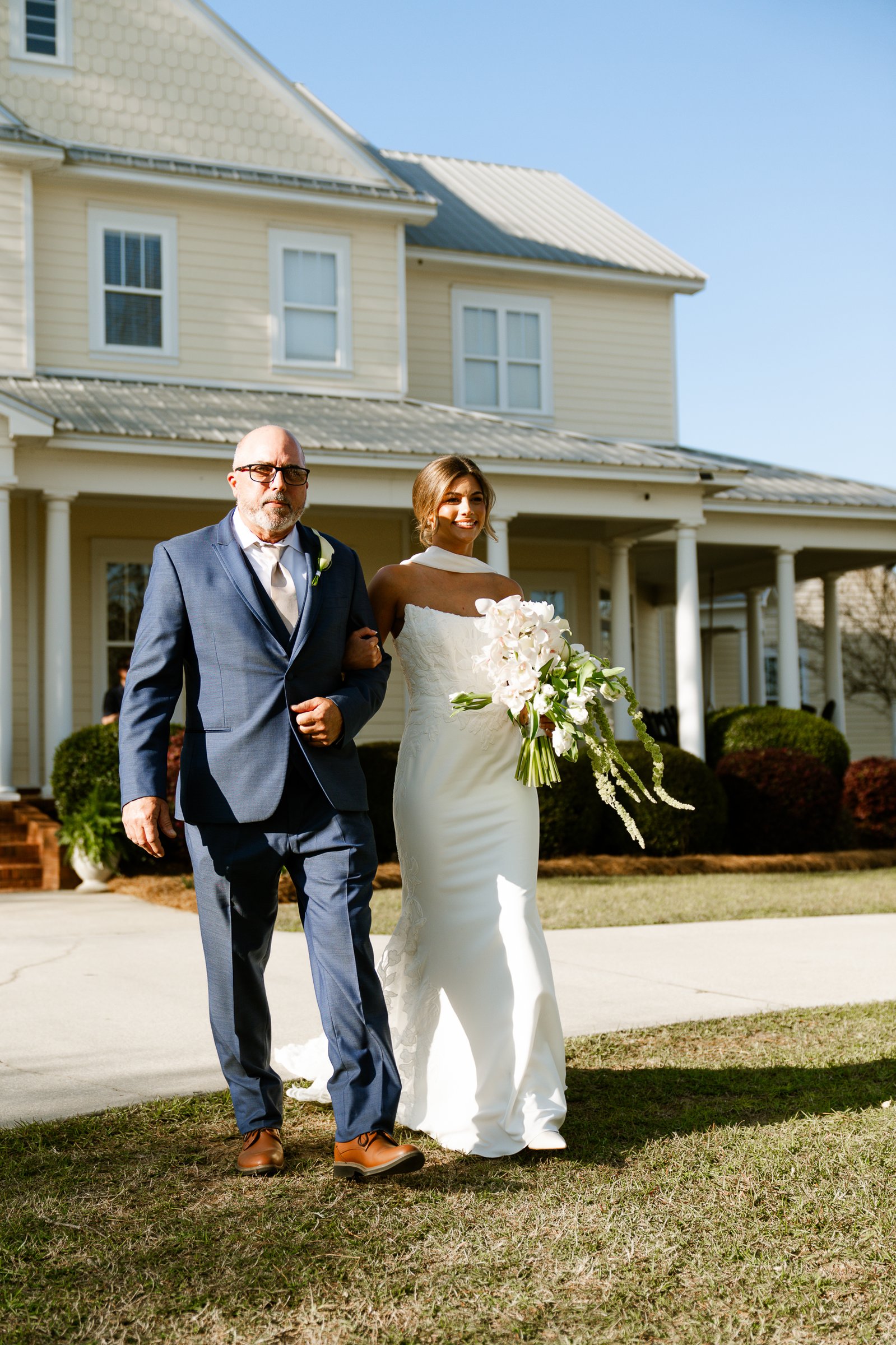 Joley carries a white bouquet and walks arm in arm with a man in a blue suit across a lawn in front of a yellow house.