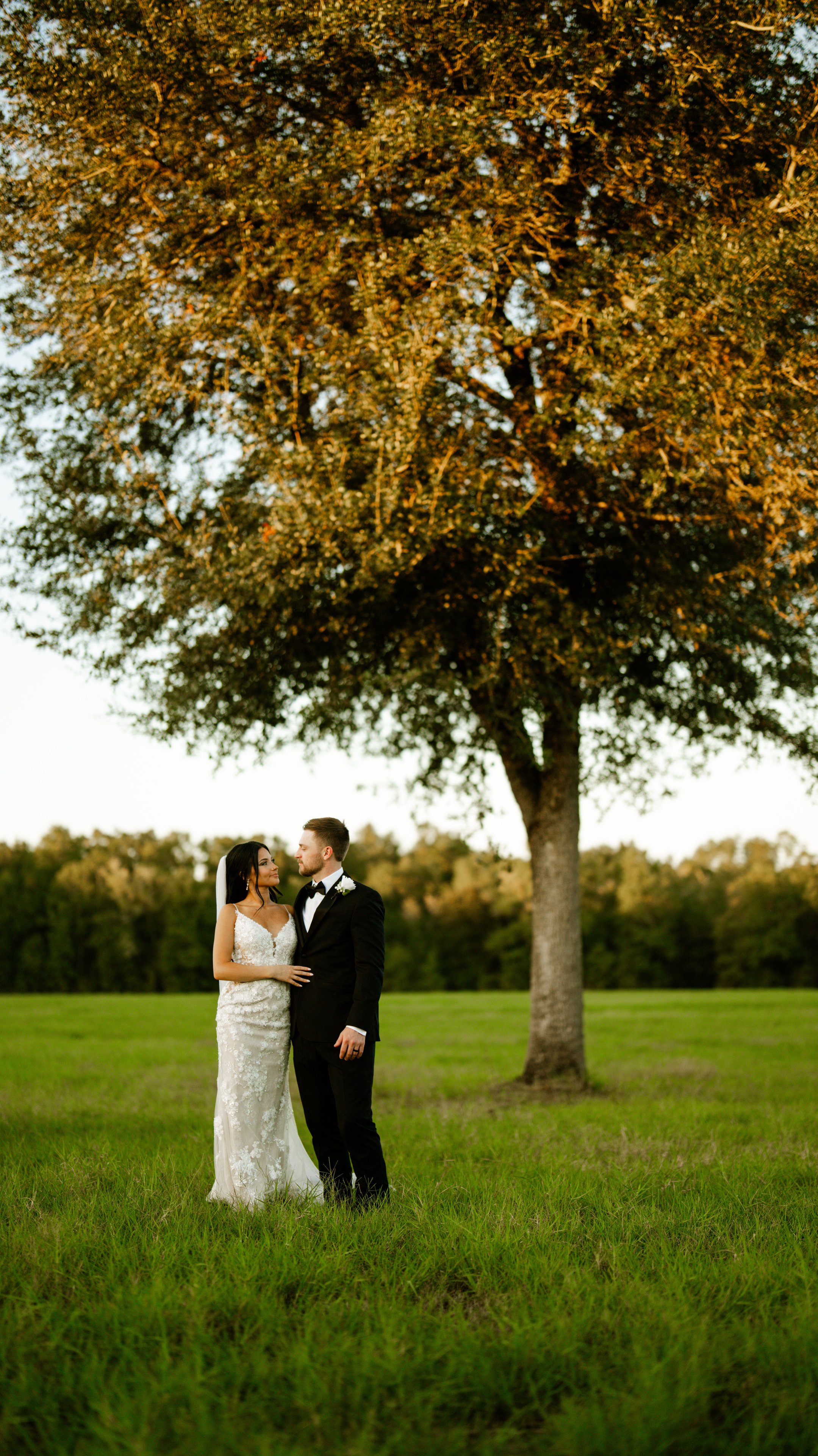 Kenadee and Corey standing close together in a green field in front of a large tree.
