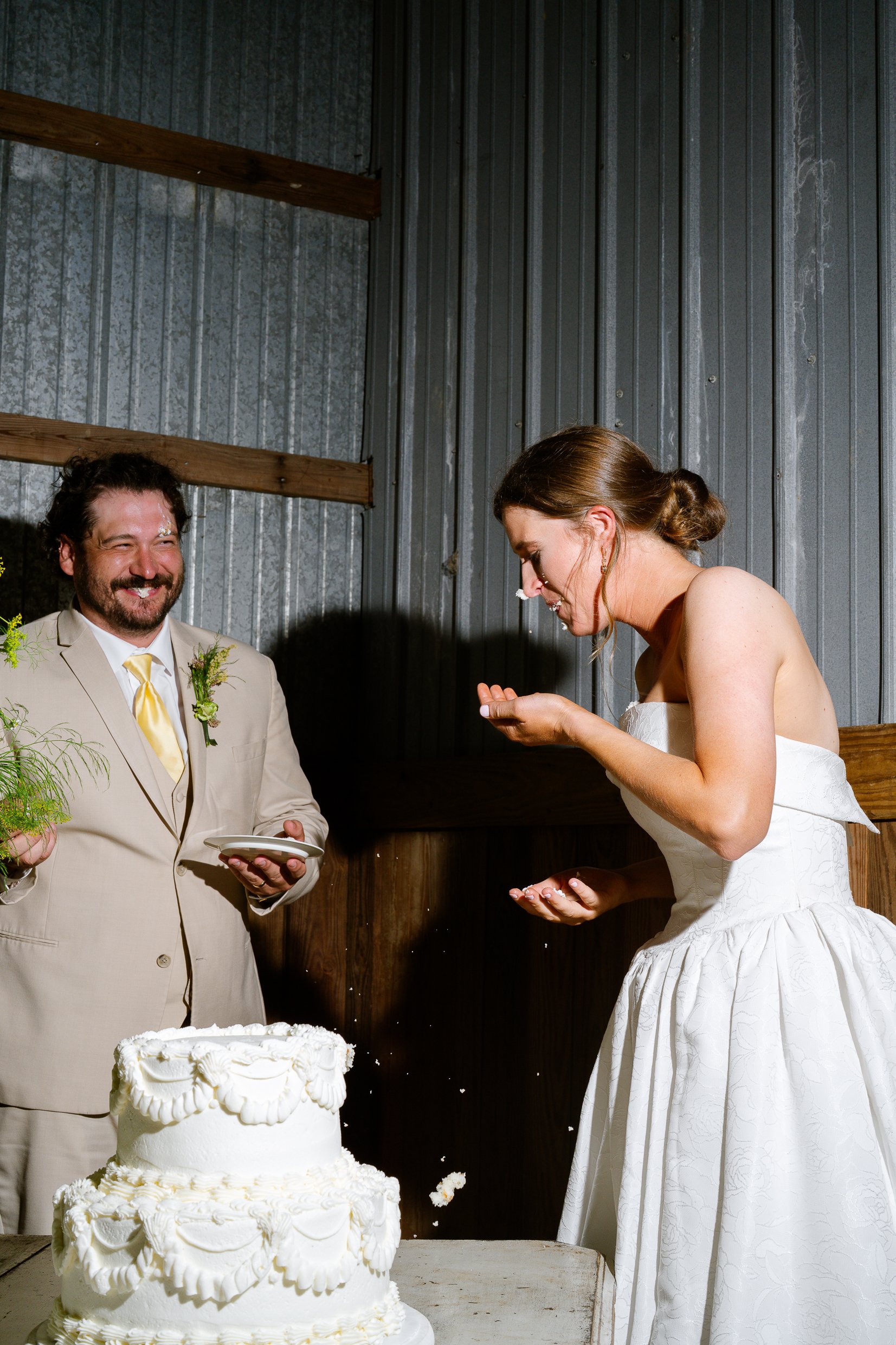 Dustin smiles and looks on while Elizabeth wipes her hands after the cake, in front of the barn wall.