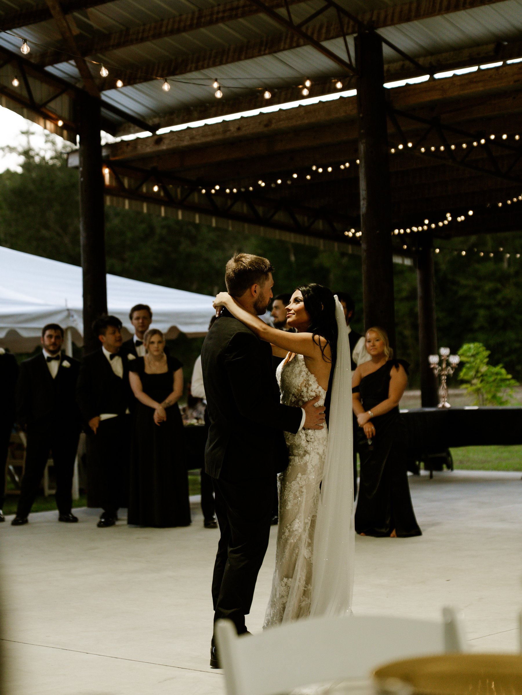 Kenadee and Corey dancing under a pavilion strung with lights while guests in dark clothes watch from the side.