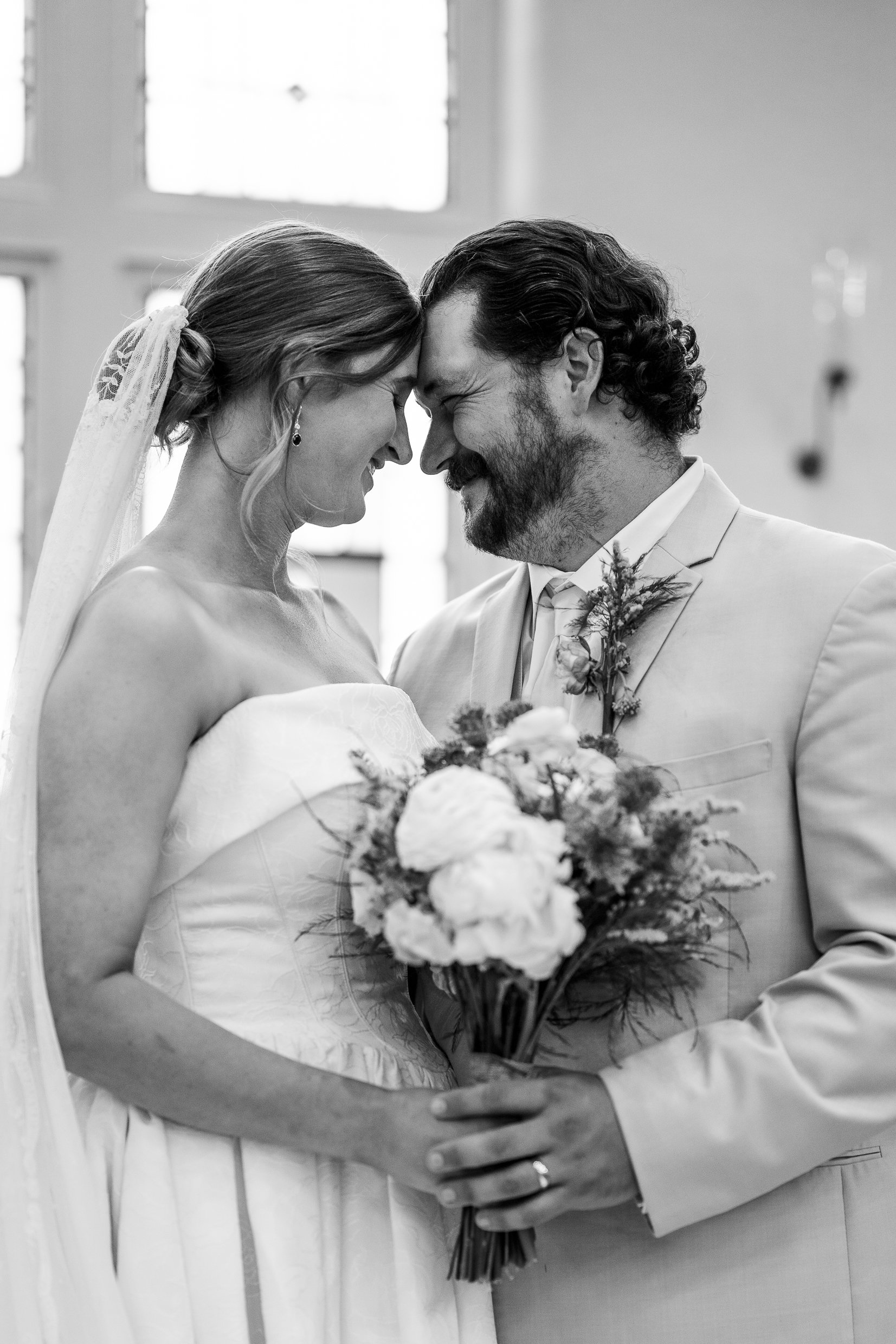 In black and white, Elizabeth and Dustin touch foreheads in front of church windows, her bouquet between them.