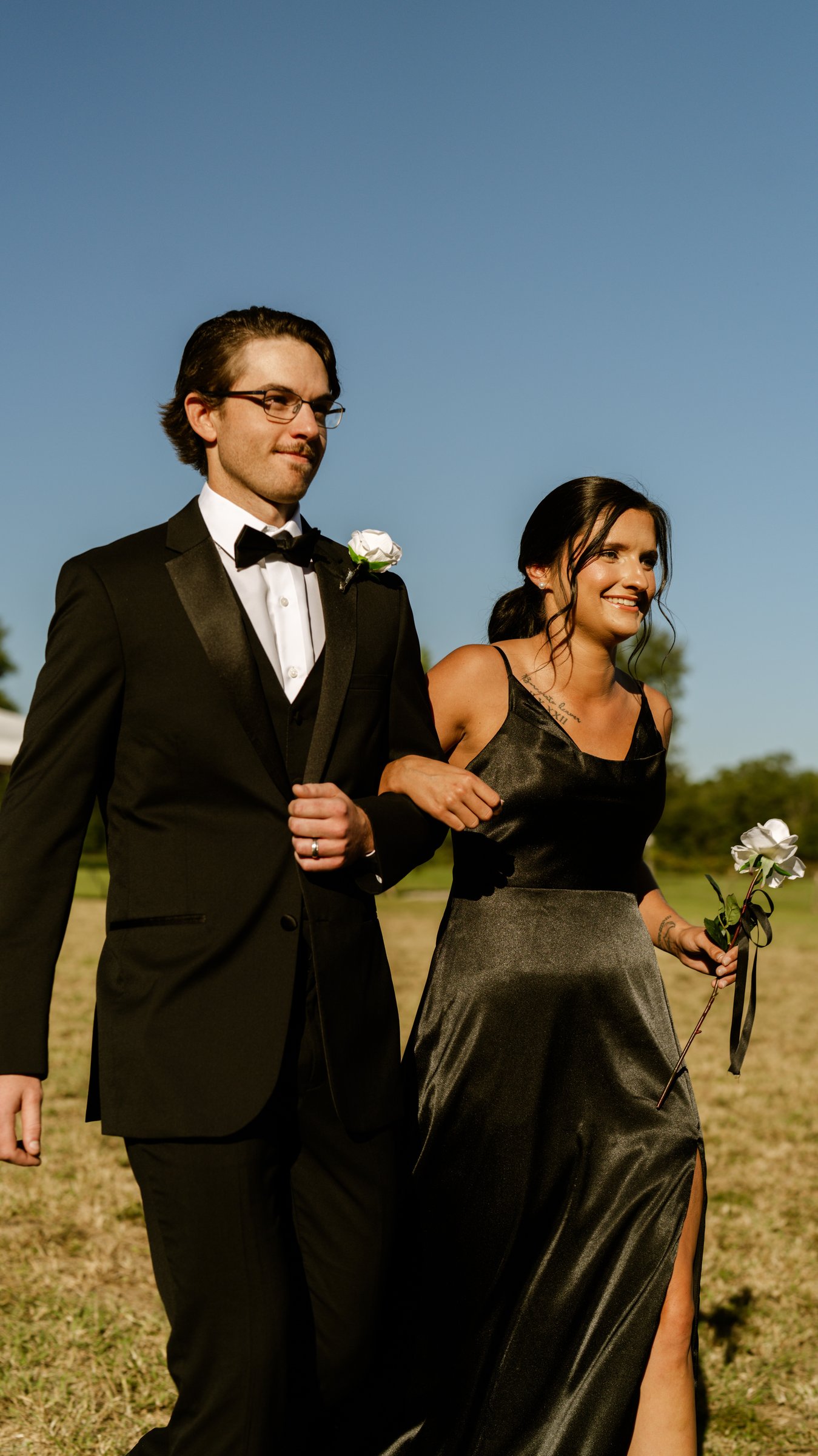 A man in a tuxedo and glasses walks arm in arm with a woman in a long black dress, holding a white flower.