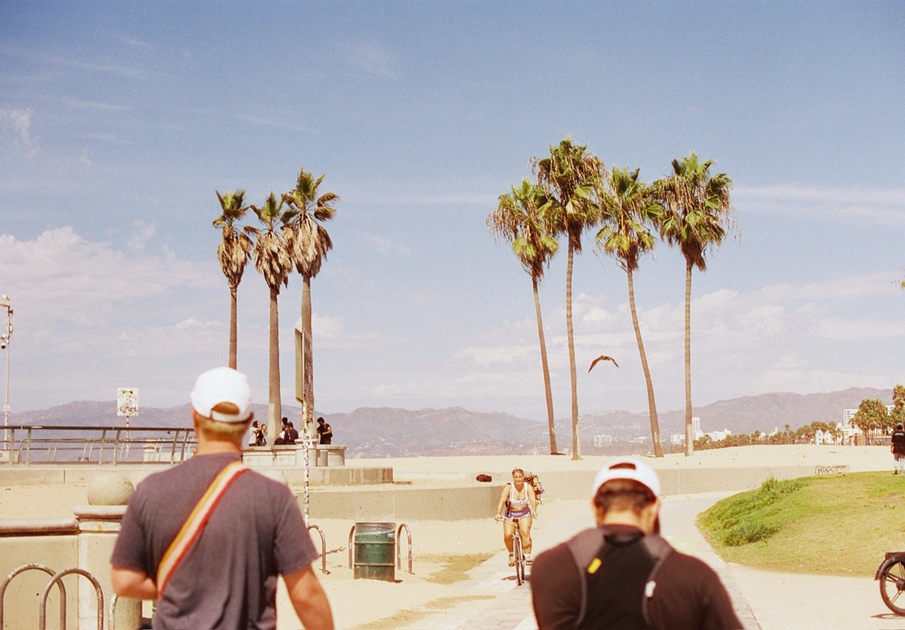 Two Midsummer crew members on the Venice Beach boardwalk beneath palm trees, photographed on film during the California wedding trip.