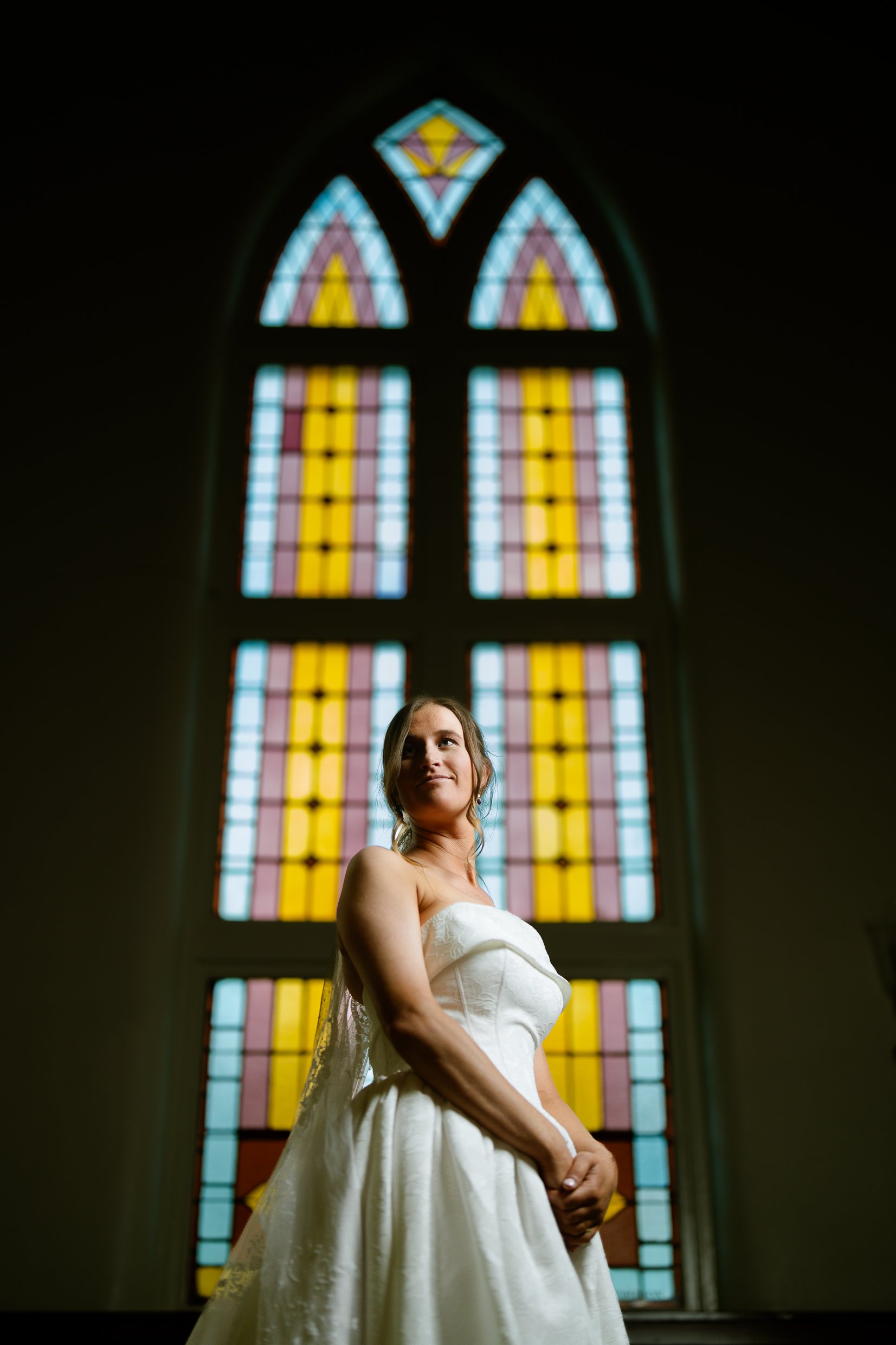 Elizabeth stands in front of a tall stained glass window of blue, pink and yellow panes, looking up and away with a slight smile.