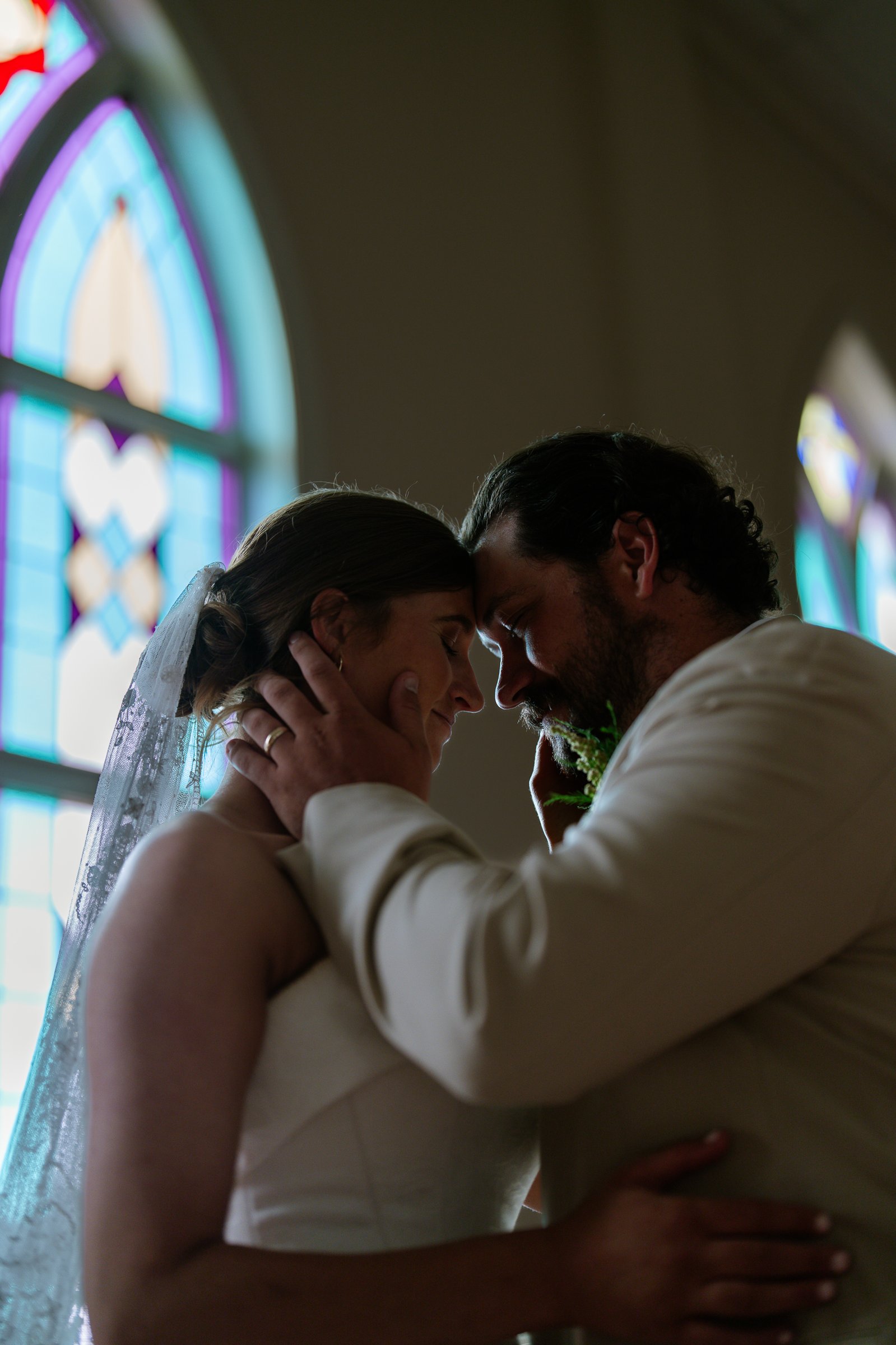 Elizabeth and Dustin touch foreheads with a stained glass window behind them, her veil and one hand against his face.