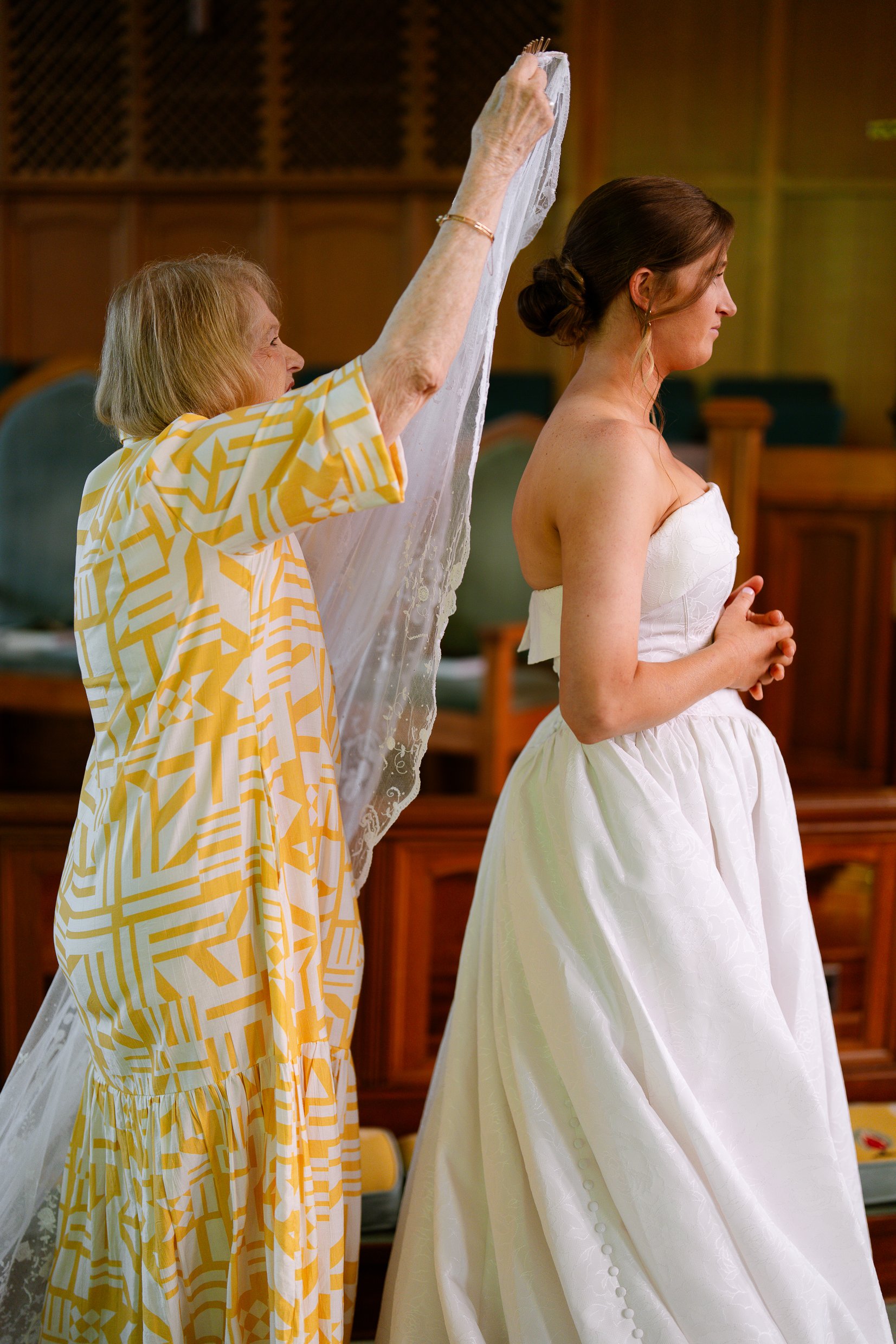 A woman in a yellow patterned dress lifts Elizabeth's long lace veil as Elizabeth stands with her hands clasped.