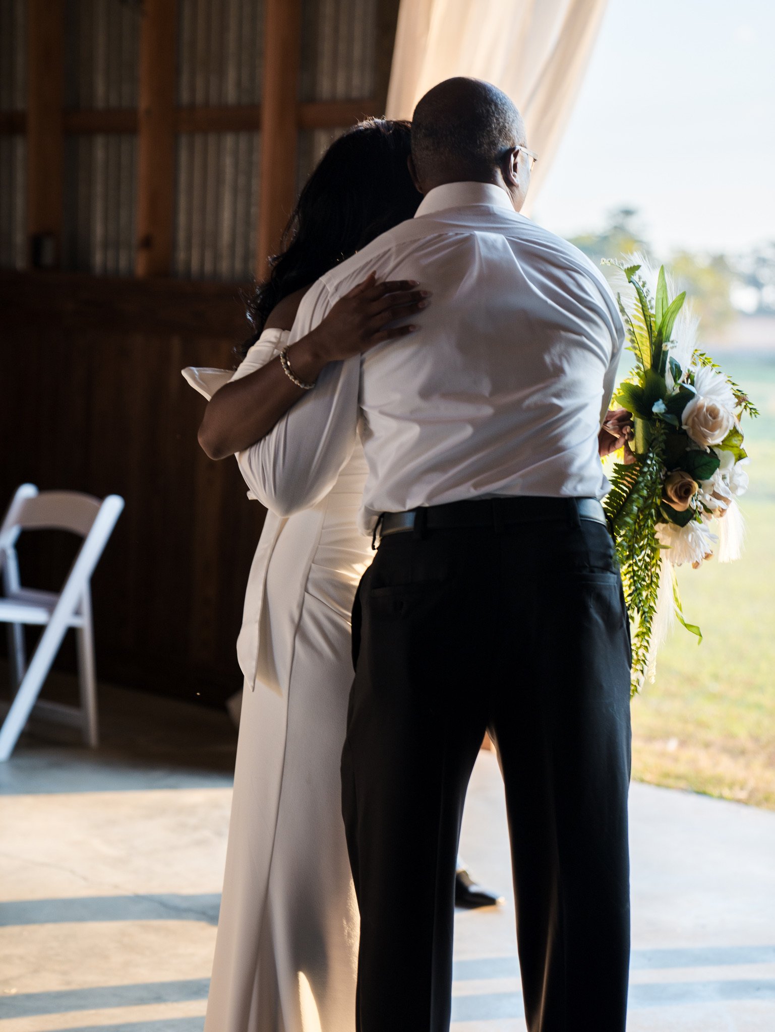 Ashley hugs a man in a white shirt in the barn doorway, with a bouquet beside them and a field behind.