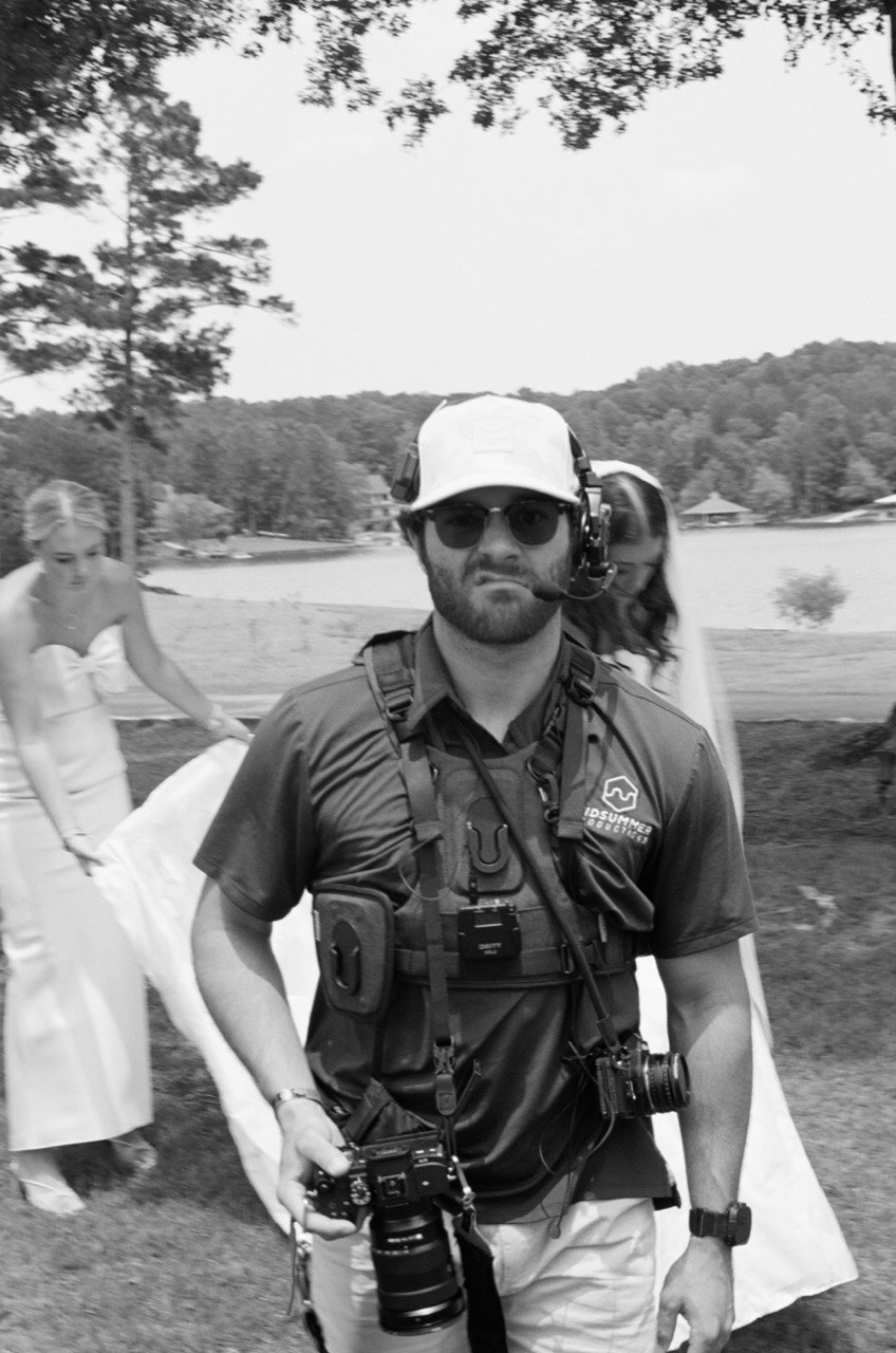 A member of the Midsummer crew wearing headphones and a camera harness, with Brooklyn behind him at New Water Farms, photographed on black-and-white film.