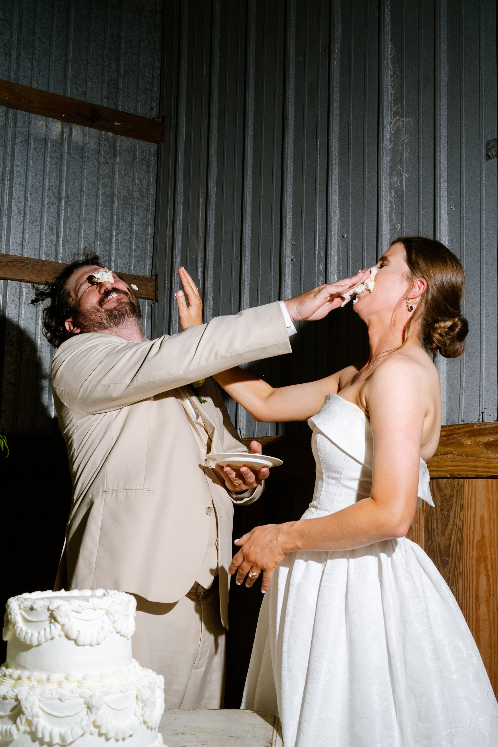 Elizabeth presses cake toward Dustin's face as he leans back laughing, a plate in his hand and the white cake beside them.