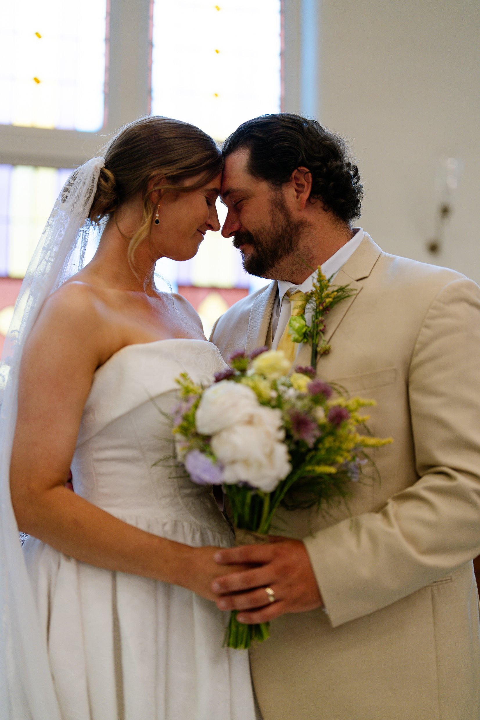 Elizabeth and Dustin touch foreheads in front of church windows, her bouquet of white and lavender flowers held between them.