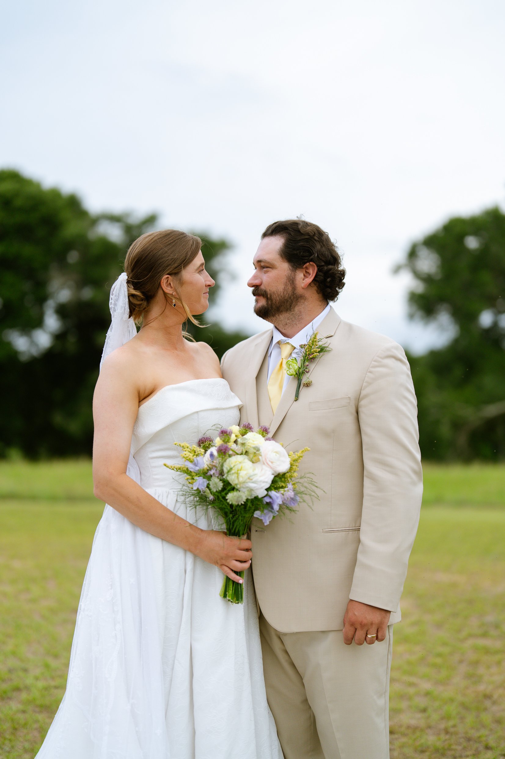 Elizabeth and Dustin stand facing each other on a grass field with trees behind them, she holds a bouquet of white and lavender flowers.