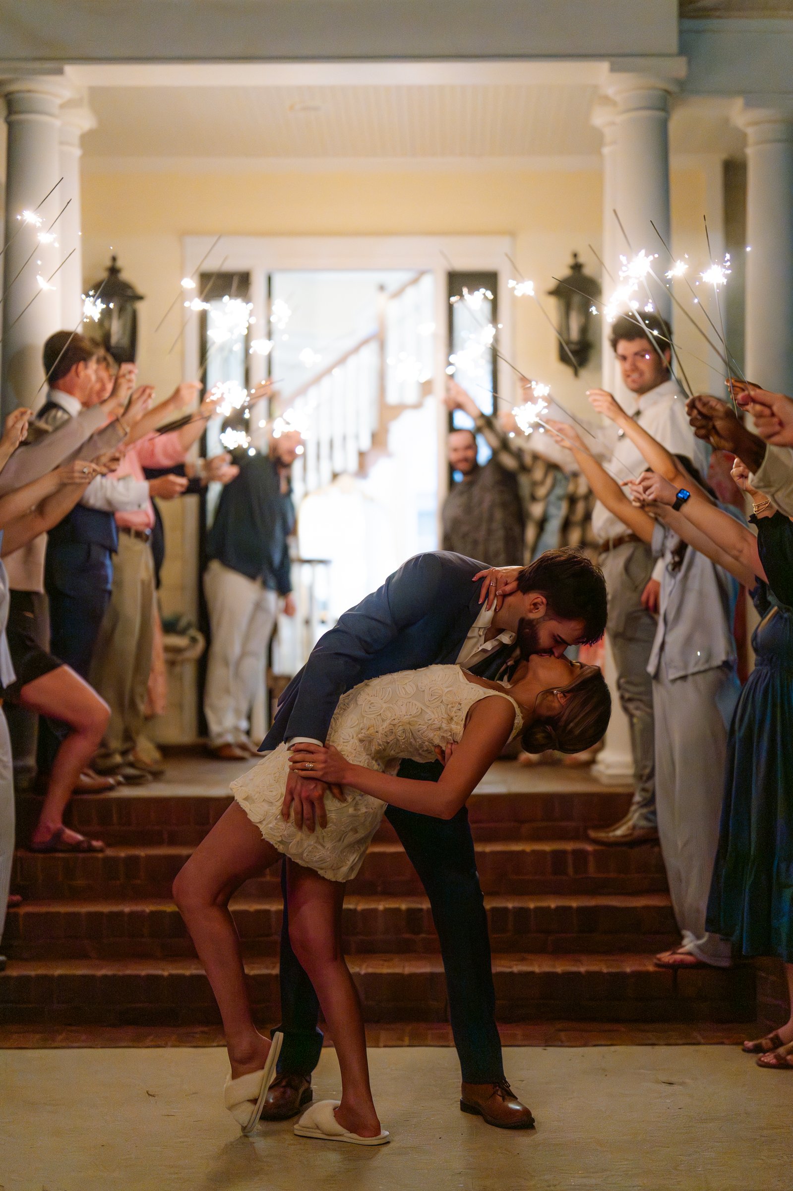 Joley, in a short white dress, is dipped backward by Matthew in a kiss on porch steps between two rows of guests holding sparklers.